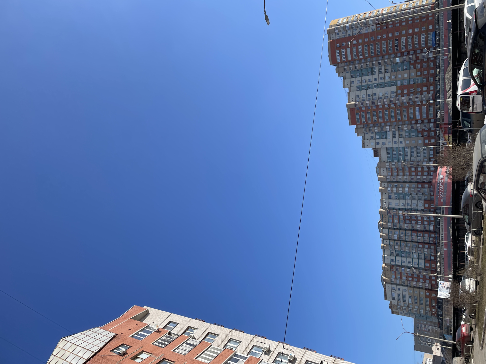 The image shows a view upwards towards the sky in Izhevsk, Russia, with two buildings visible on opposite sides of the frame.
On the bottom left, a lower building with red brick and white accents can be seen. It has multiple windows, some with air conditioning units.
On the top right, a much taller apartment building stands, displaying a red and white color scheme. Its facade is dense with numerous windows.
Between the buildings, a clear blue sky is visible. Power lines stretch across the sky. The building on the right has a sign with "AUTOMOTIV" written on it. A few cars are visible to the right of the taller building.