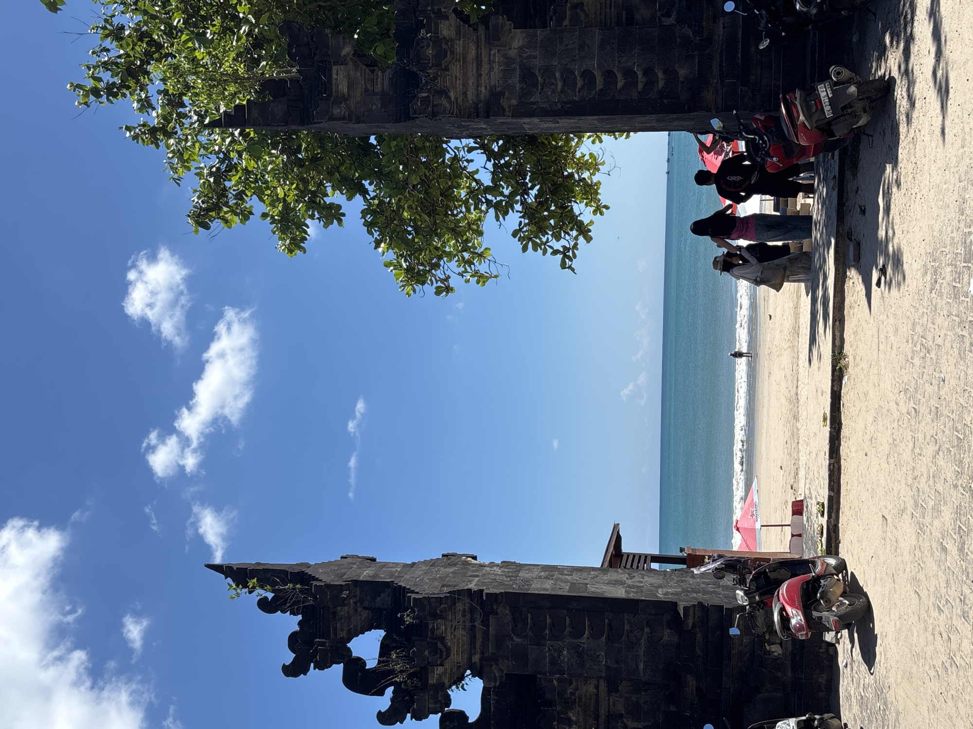 The image captures a view of the beach in Kuta Selatan, Indonesia, framed by two structures, possibly the remnants of an old gate or wall. The sky is a clear blue with scattered clouds, and a tree with green leaves extends into the frame. People stand on the beach, near the shore, facing the ocean. Some are closer to the viewer, while others are further away. Motorcycles are parked near the beach on the paved area in the foreground. A red umbrella stands on the beach, near the people and ocean.