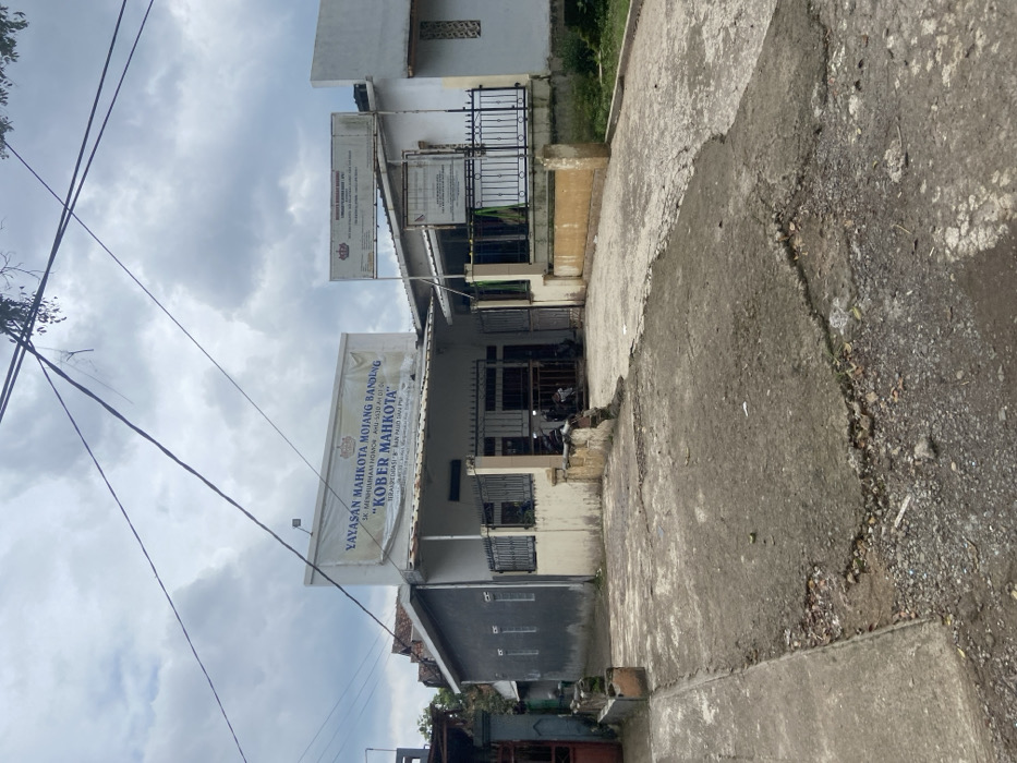 The image shows the front of a building in Ciparay, Indonesia, located at coordinates -7.022 latitude and 107.679 longitude. The building appears to be a small, single-story structure with a grey exterior. There are two motorcycles parked in front of the building. A sign above the entrance reads "YAYASAN MAHKOTA MOJANG BANDUNG," with a smaller sign beneath it that says "KOBER MAHKOTA." There is another sign on a post near the building, but the text is unreadable. The building is surrounded by a black metal fence and is situated on a dirt driveway. The driveway is uneven and cracked, and there is vegetation growing in the cracks. The sky is cloudy, but there is no indication of rain.