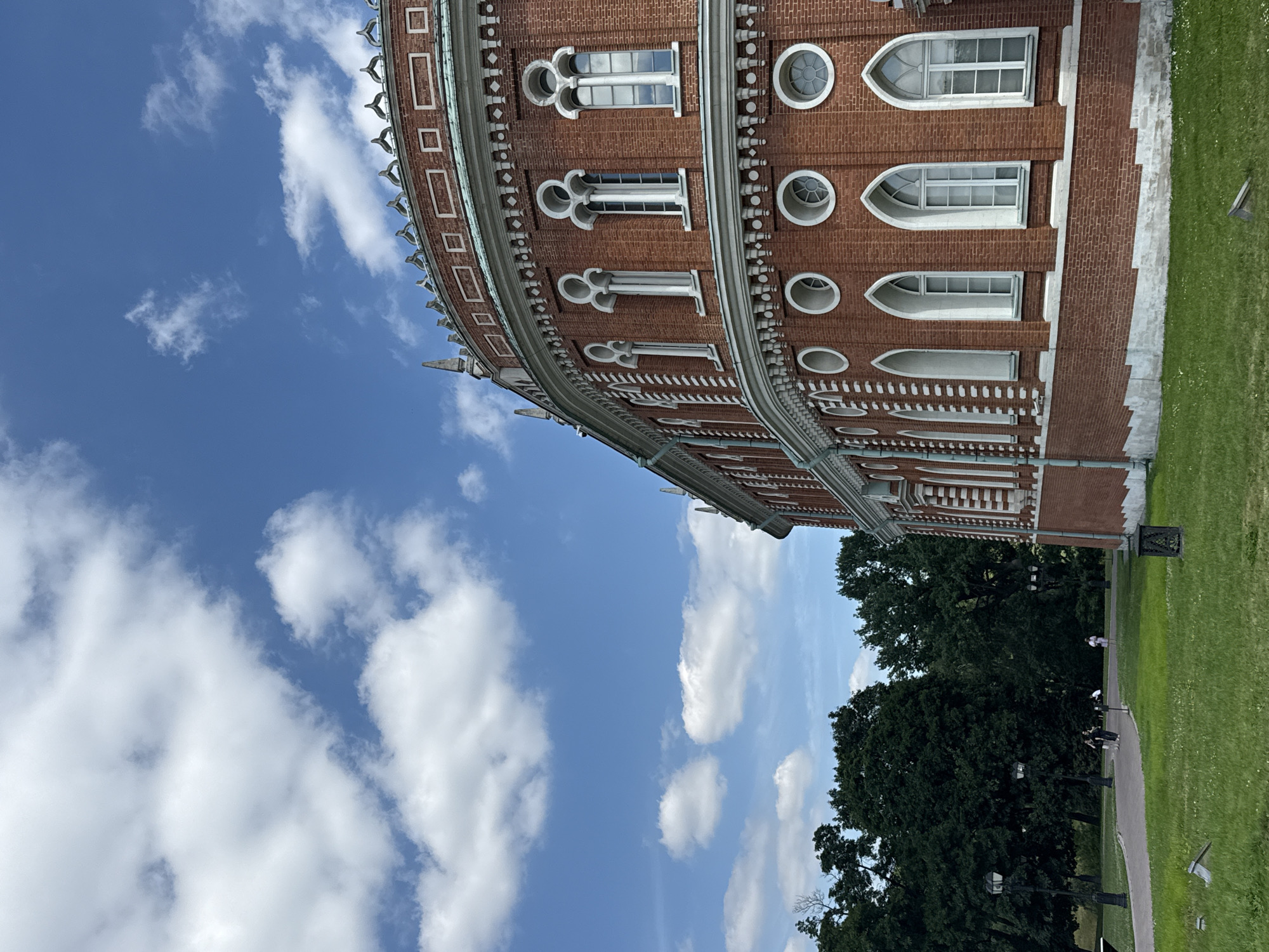 This media file captures a close-up, low-angle shot of a red brick building with ornate white trim and various window shapes, including arched, round, and rectangular openings. The building's curved facade is visible, suggesting a semicircular or circular structure. Above the building, a bright blue sky is filled with fluffy white clouds, indicating fair weather during the daytime. To the right of the building, a vibrant green lawn stretches out, with a paved pathway and several trees forming a backdrop. A few distant figures can be seen on the pathway, walking or standing. The overall scene depicts an architectural landmark in a park-like setting, bathed in natural light. The time of day appears to be midday or early afternoon. No visible text is present in the image. The architectural style and the presence of lush greenery suggest a historical park or estate, consistent with locations in Moscow, Russia.