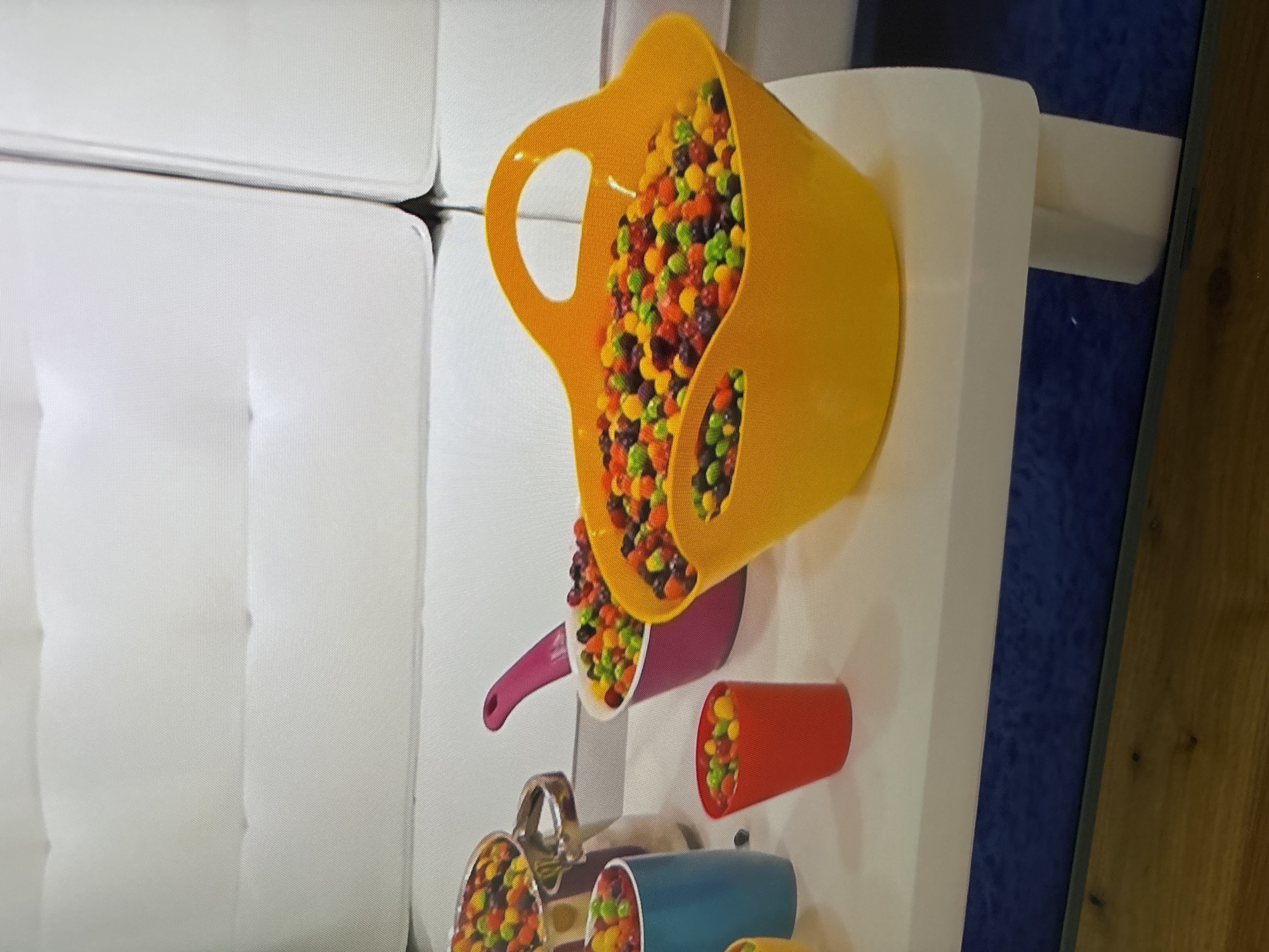 A close-up shot shows a collection of brightly colored candies filling various containers arranged on a white table. The containers include a silver pot, a pink bowl with a handle, a blue cylindrical cup, a red cup, and a large yellow basket with handles. The candies themselves are small and oval-shaped, exhibiting a vibrant mix of colors such as red, orange, yellow, green, purple, and blue. The background features a section of a white sofa. The scene does not provide any information about the city of Dubai, United Arab Emirates.