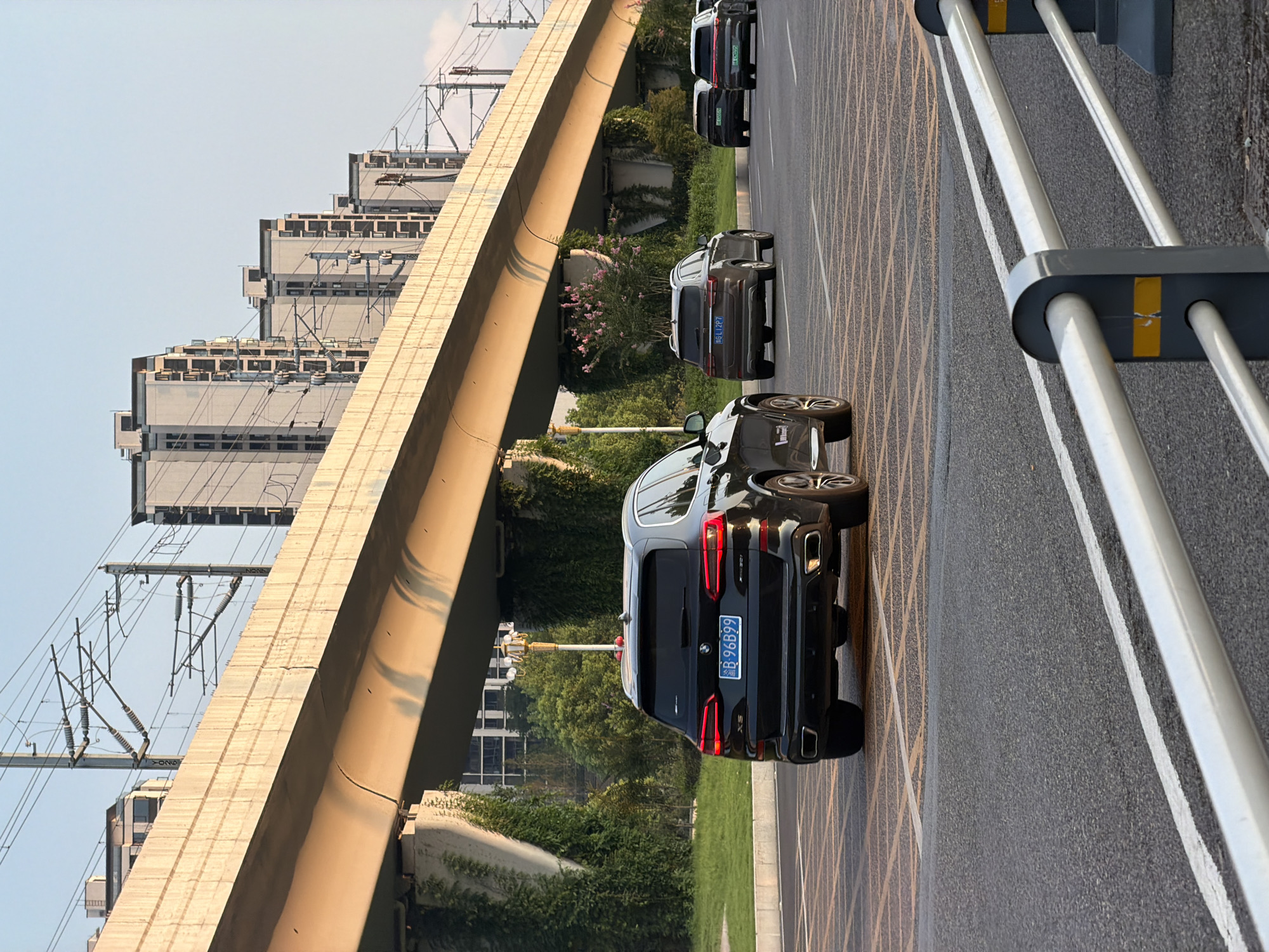 The image depicts a street scene in Jiangjin, China, under a large, elevated overpass that appears to be part of a transportation infrastructure, possibly for trains due to the overhead electrical lines. The overpass casts a warm, golden glow on the road below, likely from the setting sun. Several vehicles are driving on the road. In the foreground, a black BMW SUV with license plate "B 96B99" is prominently featured. Further down the road, a brown SUV with license plate "蒙L12P7" is visible, followed by two dark-colored cars, one of which has a license plate that appears to be "京E C005" or similar. The surrounding environment includes lush green vegetation growing on the pillars supporting the overpass, and in the background, several multi-story residential buildings are lined up against a clear sky. The overall impression is a snapshot of urban life during the golden hour.