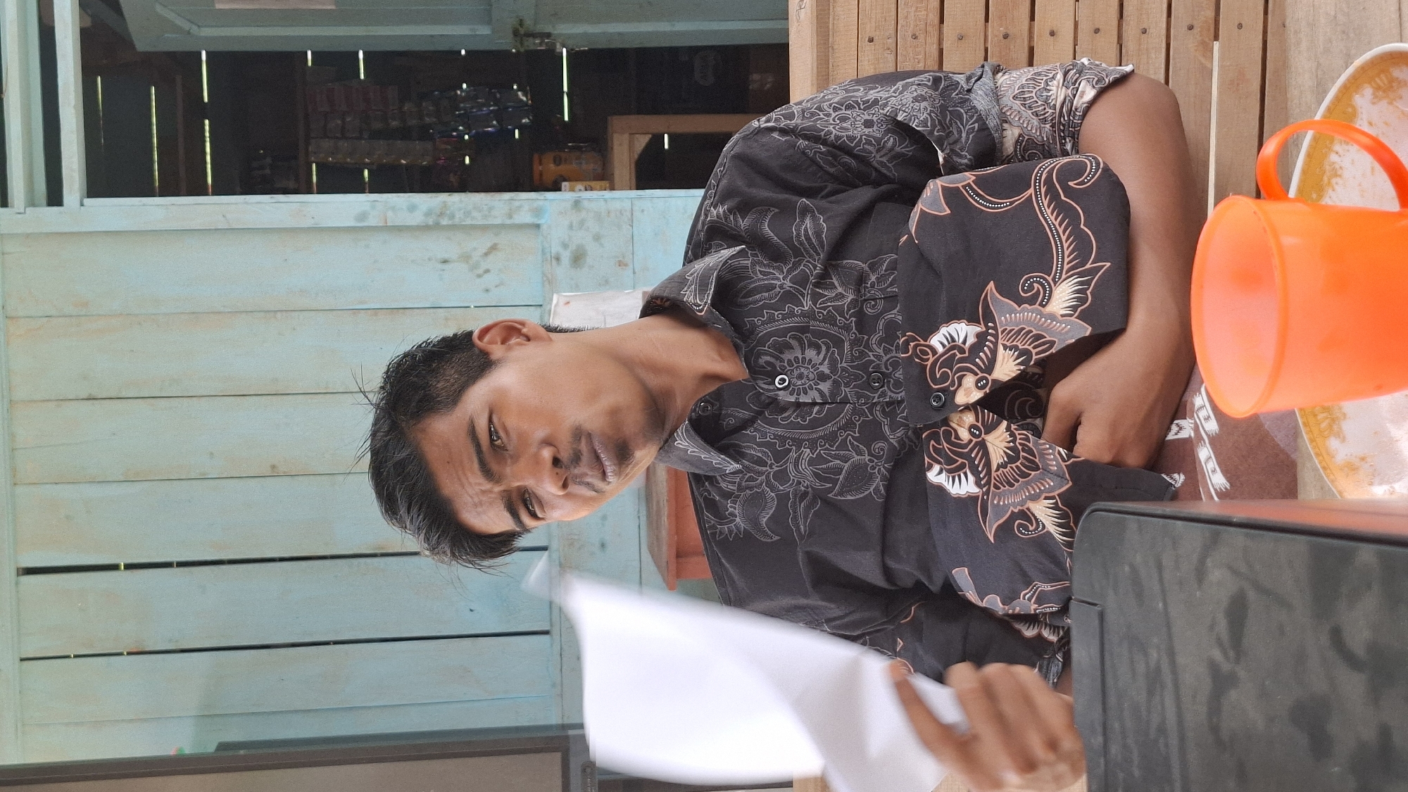The image depicts a medium shot of a man sitting on a wooden bench in front of a light blue wooden wall. The man is wearing a black patterned shirt. He is holding a piece of white paper in his left hand and looking towards the viewer with a slightly raised eyebrow and pursed lips. An orange cup is visible on a table in the foreground, to the man's right. The background shows a doorway leading to a room with shelves and various items. The setting appears to be an outdoor or semi-outdoor area, possibly in Grong-grong, Indonesia.