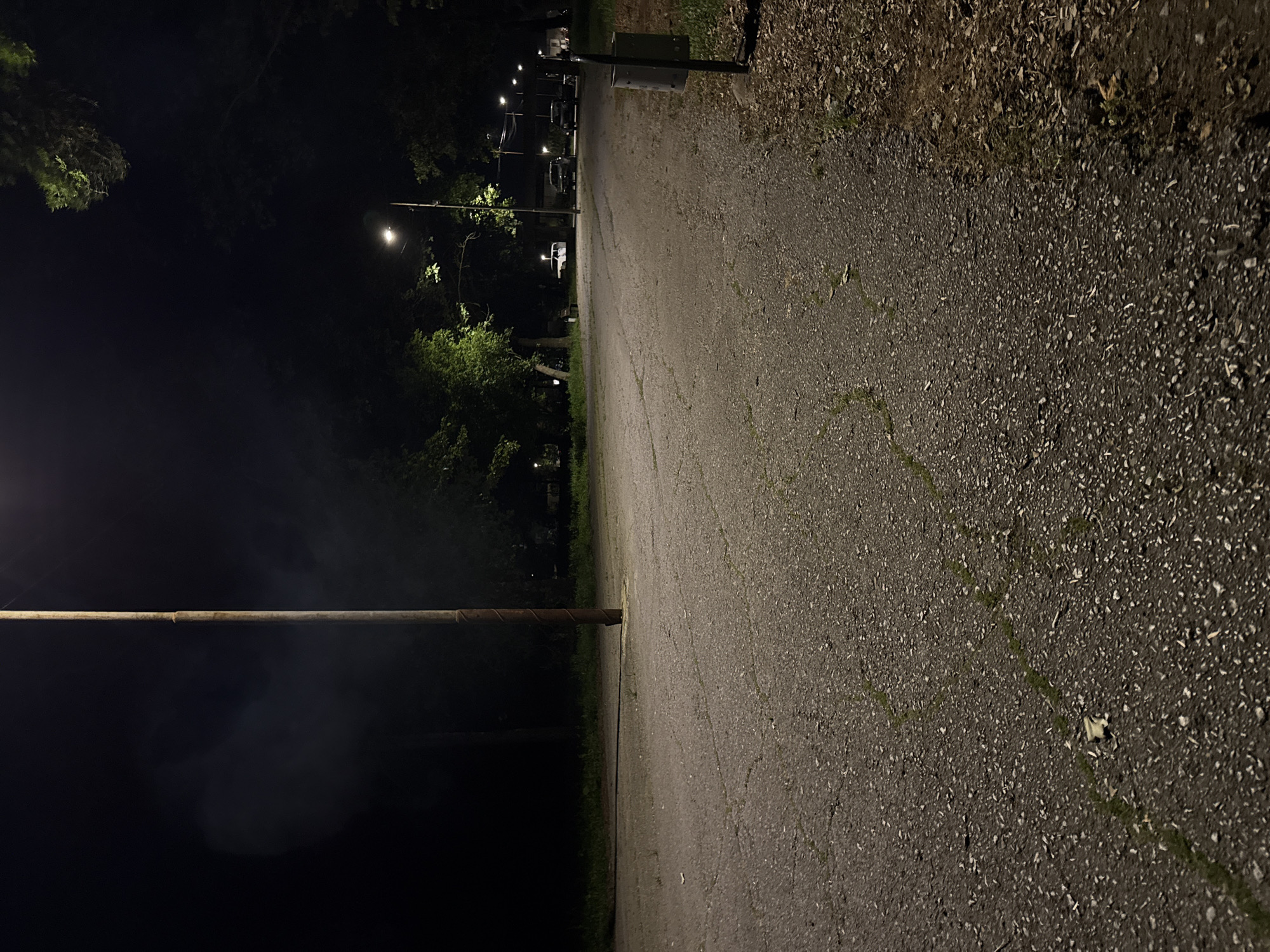 The image depicts a dimly lit asphalt road at night. A wooden pole is visible in the left foreground. The road curves slightly and is lined with trees on the left side and foliage on the right. Streetlights illuminate the road, and several cars are parked in the distance, indicating a residential or recreational area. The precise location within Elhovo, Bulgaria cannot be determined without additional information.