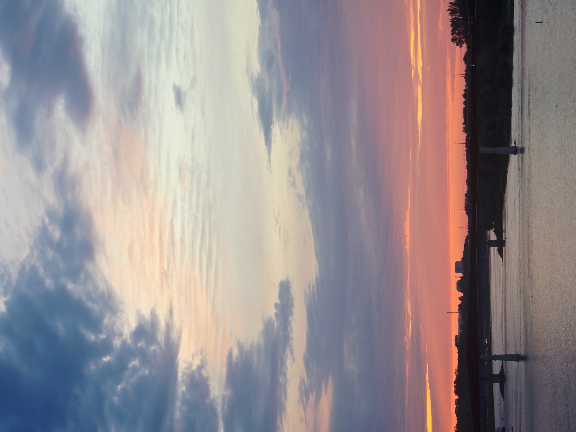 The image depicts a bridge crossing a body of water, likely the Vistula River, in Warsaw, Poland. The sky above is a vibrant mix of sunset colors, with hues of orange, pink, and purple blending into shades of blue and gray clouds. The bridge itself is a prominent structure, appearing to be a road bridge with a dark red railing. Several concrete pillars support the bridge, extending into the water. In the background, a treeline borders the far bank of the river, and beyond that, city buildings and industrial structures are visible. The water reflects the colorful sky, creating a shimmering effect on its surface. There are no people visible in the image.