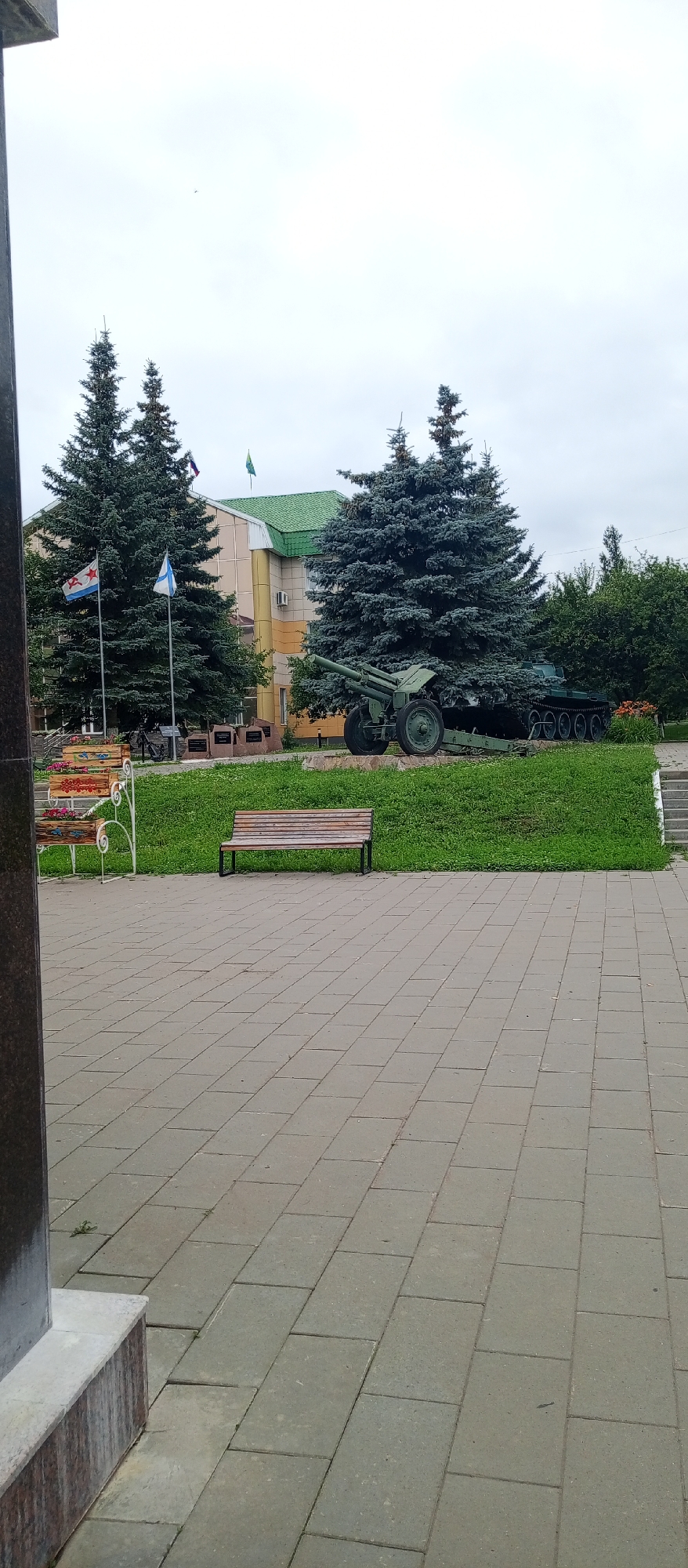 The image shows an outdoor scene with a building in the background, surrounded by trees and a paved area. In front of the building, there are several large evergreen trees. A military exhibit is displayed on a grassy area between the trees and the paved walkway. This exhibit includes what appears to be a cannon and a tank. Several flags are flying from poles in front of the building.
To the left of the exhibit, there is a colorful bench. A plain wooden bench is situated on the grassy area closer to the foreground. The ground in the foreground is paved with grey rectangular tiles. The sky is overcast, suggesting a cloudy day. There is no clear indication of the time of day. The overall setting appears to be a public space, possibly a memorial park or a historical site, given the military equipment and flags. No people are visible in the frame. There is no discernible text in the image. The scene is static, and no activity is taking place.