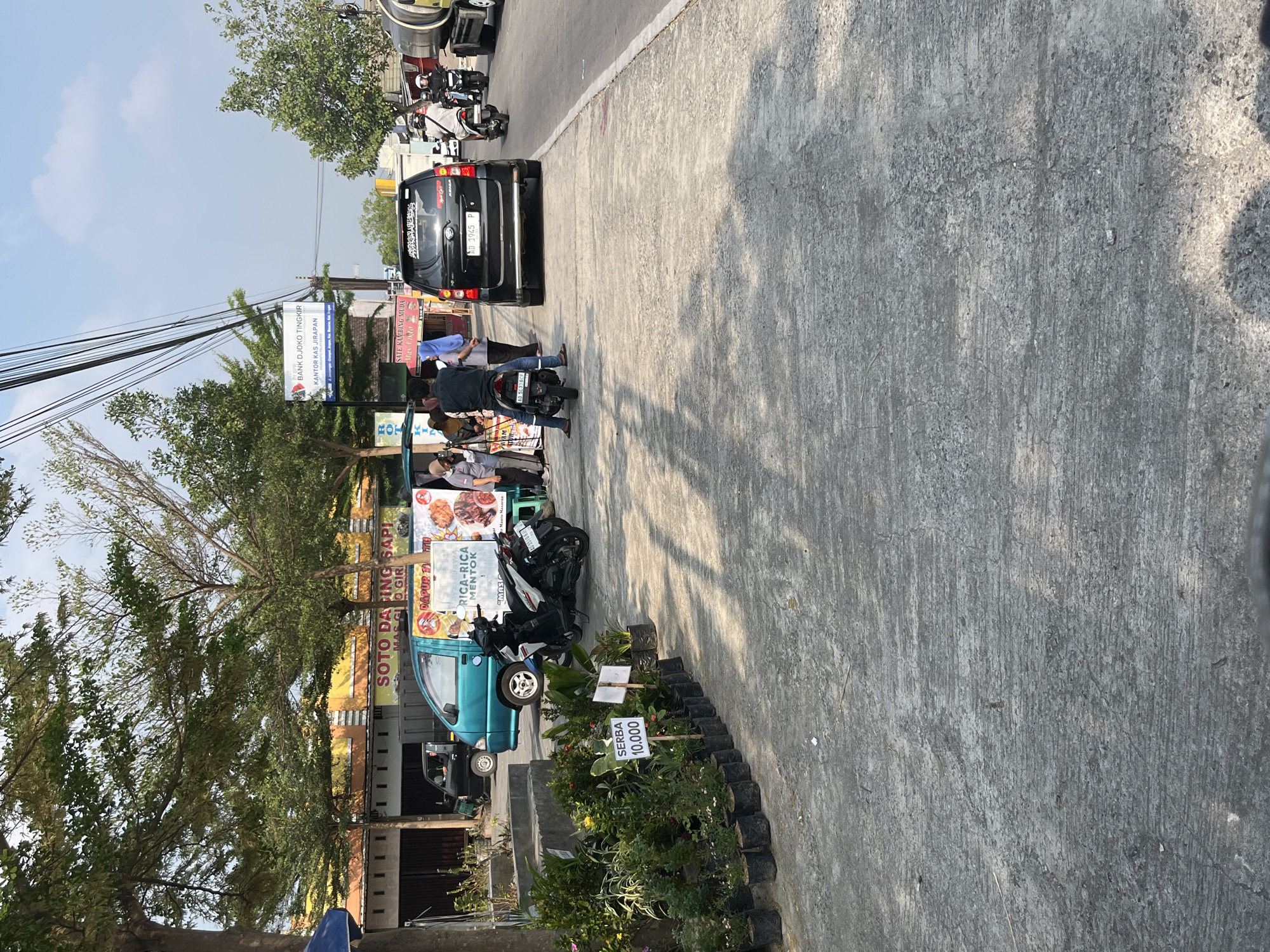 The image depicts a street scene in Munggur, Indonesia, on a sunny day with a clear blue sky and scattered clouds. The foreground is dominated by a concrete sidewalk and road. On the left side of the sidewalk, a row of potted plants is displayed for sale, with a sign indicating "SERBA 10.000" (Everything 10,000).

Further down the street, several vehicles are present. A teal-colored car is parked, and next to it, a food stall is set up with signs advertising "SOTO DAGING SAPI" (Beef Soto) and "RICA-RICA MENTOK" (Duck Rica-Rica). A few people are gathered around the stall, seemingly interacting with vendors. A white and blue motorbike is parked near the stall, and another motorbike with a rider is visible behind it.

A black SUV is parked on the right side of the road, with a license plate clearly visible. Further down the road, a large tanker truck is driving away from the camera. In the background, buildings line the street, some with colorful awnings. A sign for "BANK DJOKO TINGKIR" is also visible. The environment appears to be a typical urban or semi-urban setting with a mix of commercial activity and residential buildings. Power lines are strung across the top of the image.