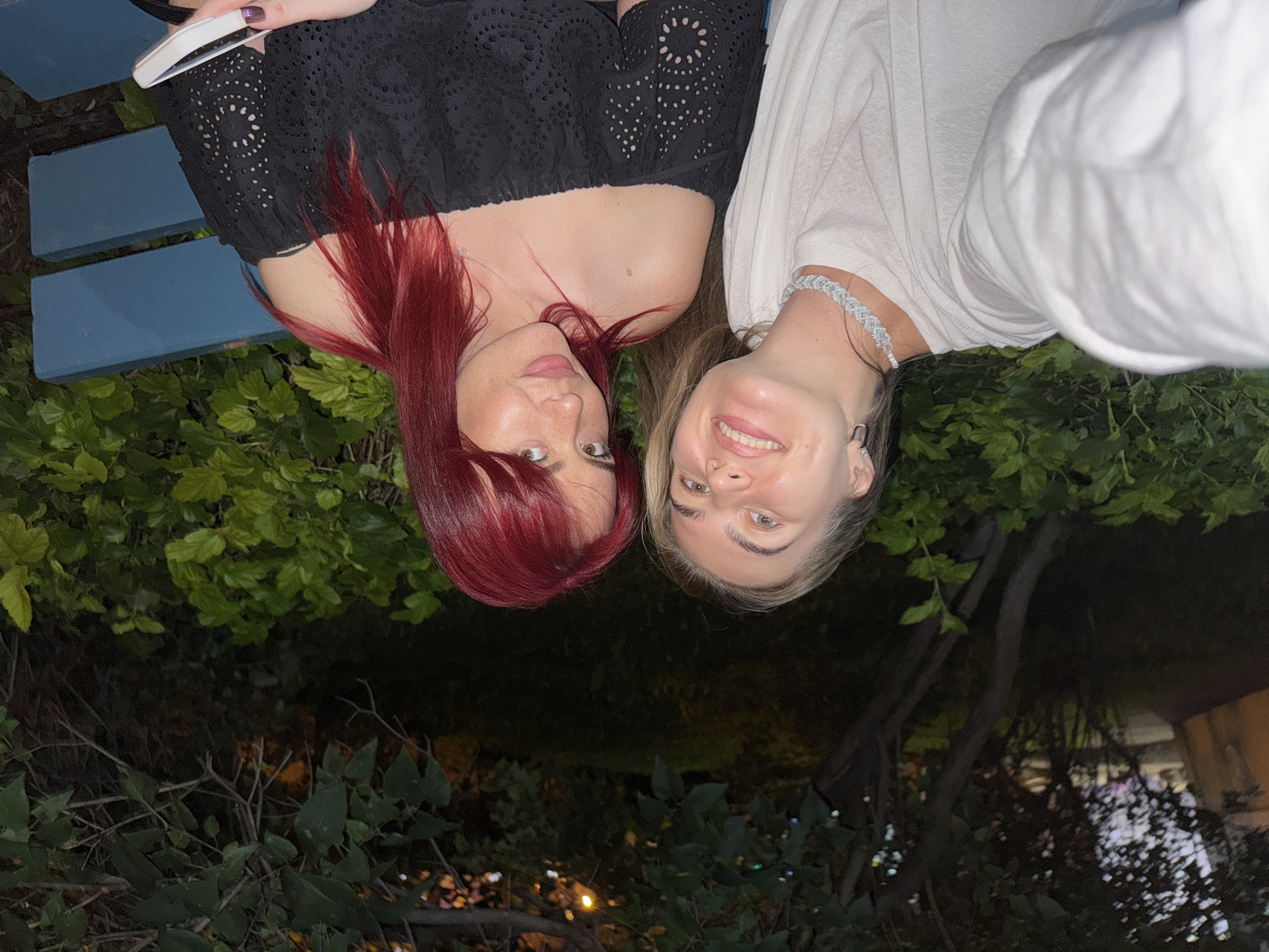 Two women are seated on a blue park bench outdoors at night. The woman on the left has blonde hair and is wearing a white shirt and a sparkly necklace. The woman on the right has bright red hair and is wearing an off-the-shoulder black top. She is holding a white smartphone in her right hand. The background consists of lush green foliage and trees, with some faint lights visible in the distance, suggesting an urban or suburban park setting. The overall lighting indicates it is evening or night.