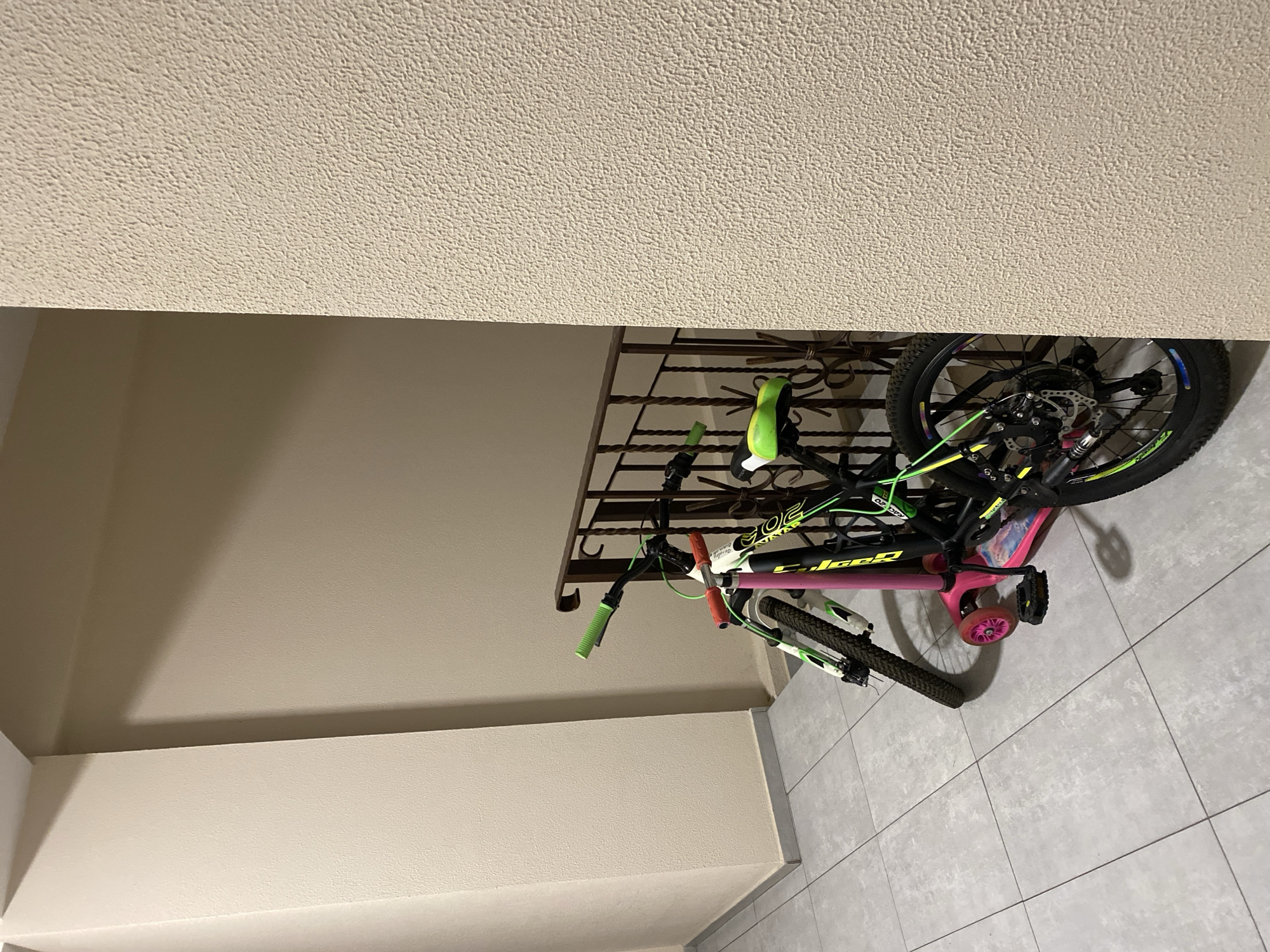 In an indoor hallway with tiled flooring, two bicycles and a scooter are parked next to a metal railing. The bicycle in the foreground is black and green with a neon green seat and handlebars. The second bicycle, partially obscured, is white and pink. A pink scooter with two wheels is positioned in front of the bicycles. The walls are painted in a neutral, textured beige. The scene appears to be in a residential building or common area. The location in Chișinău, Moldova, is not discernible from the visual information provided.