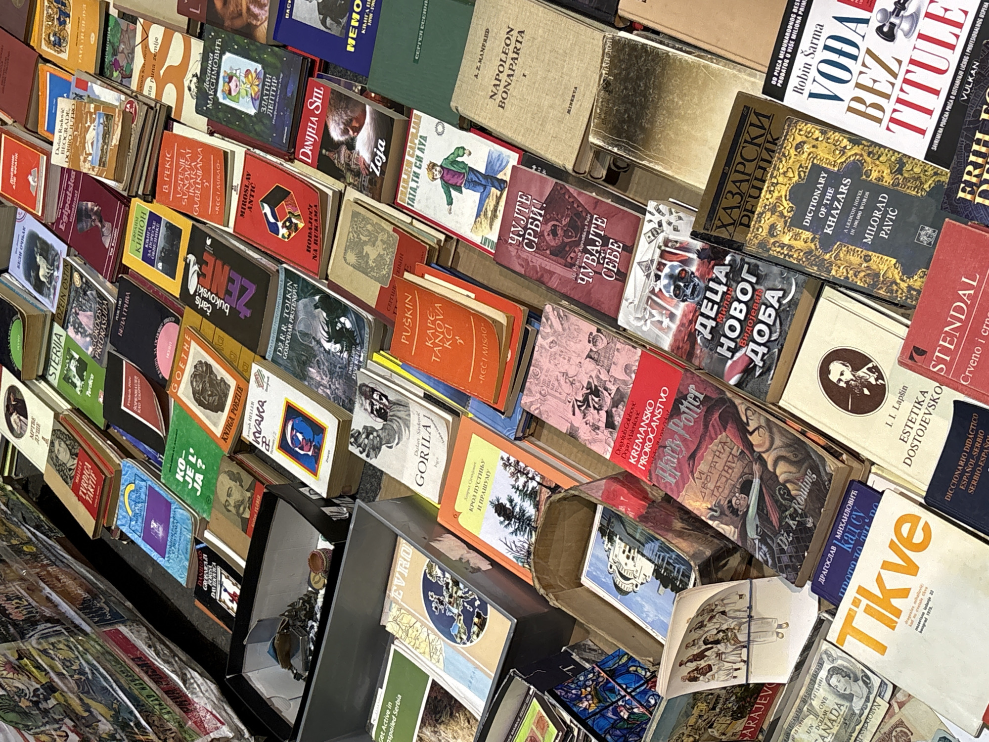 The image captures a vibrant and densely packed display of books, characteristic of a book market or second-hand stall in Belgrade, Serbia. Hundreds of books are stacked and arranged at various angles, creating an overflowing and inviting scene for browsing.
The books are diverse in genre and age, with covers visible in a mix of bright colors and more subdued tones. The dominant language is Serbian, presented in both Cyrillic and Latin scripts, though several English titles are also prominent.
Among the clearly identifiable titles and authors are Milorad Pavić's "Dictionary of the Khazars" (with both Serbian Cyrillic and English versions visible), Robin Sharma's "Vođa bez titule" (The Leader Without a Title), and several "Harry Potter" books by J.K. Rowling (Dž. K. Rouling). Other notable works include J.R.R. Tolkien's "Gospodar Prstenova" (Lord of the Rings), "Napoleon Bonaparte" by A.Z. Manfred, Charles Bukowski, Goethe, Pushkin, Miroslav Antić, Desanka Maksimović, and Jane Austen's "Gordost i predrasuda" (Pride and Prejudice). Specific Serbian titles like "Kremansko proročanstvo" (Kremna Prophecy), "Čujte Srbi! Čuvajte sebe" (Listen Serbs! Take Care of Yourselves), and "Beograde dobro jutro" (Good Morning Belgrade) further ground the scene in its Serbian context.
The bright, natural light suggests the photograph was taken during the day. In one section, a Canadian dollar bill is visible among the books, hinting at international exchange or currency used in transactions. The overall scene depicts a rich collection awaiting discovery by enthusiastic readers.