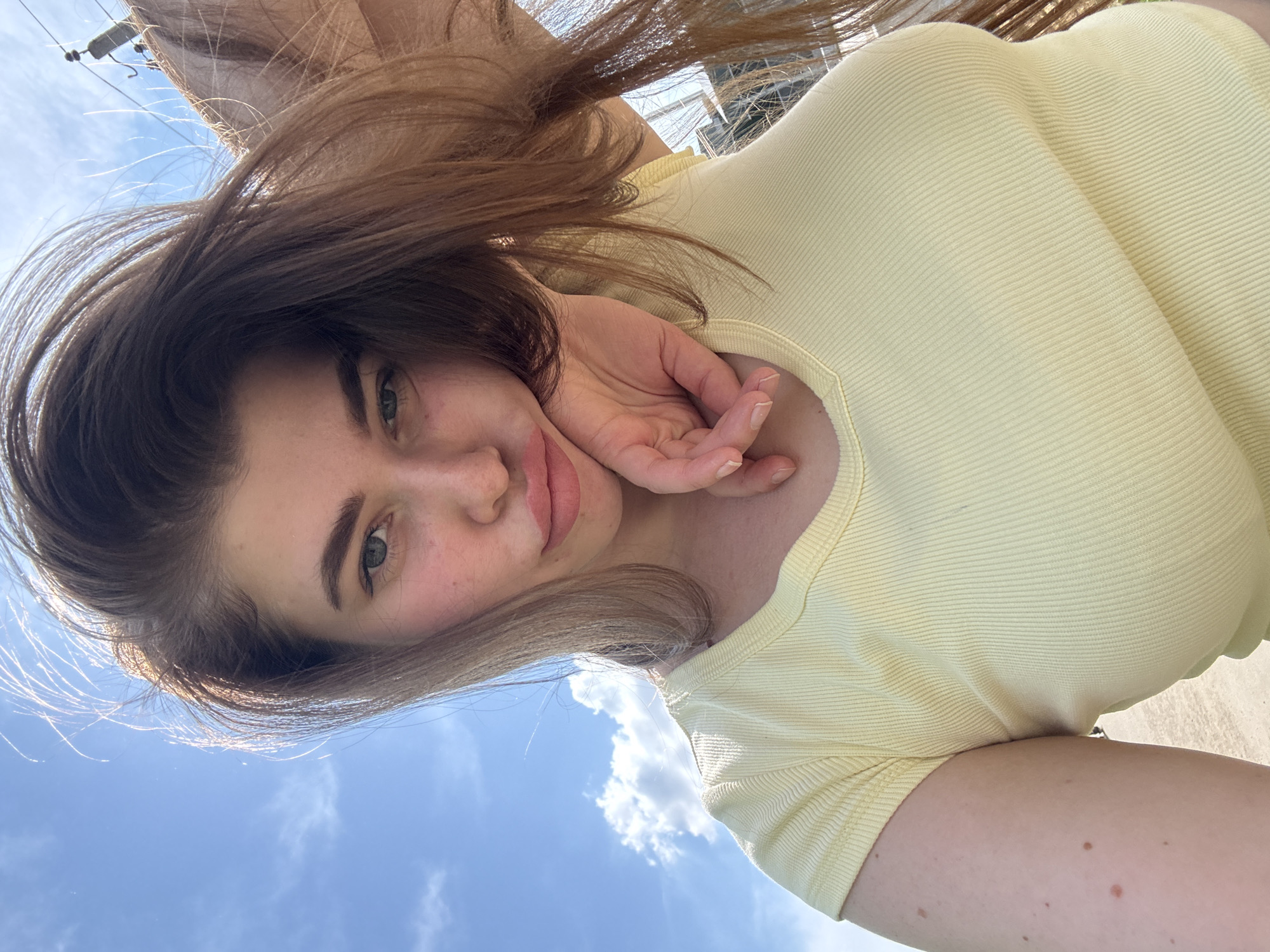 A selfie photograph depicts a woman with brown hair, fair skin, and green eyes. She is wearing a light yellow, short-sleeved, ribbed shirt. Her right hand is positioned near her face. The background consists of a clear blue sky with scattered white clouds. Electrical wires and a pole are visible at the upper left. The location is potentially Troshkovo, Russia, although there are no identifying landmarks to definitively confirm this.