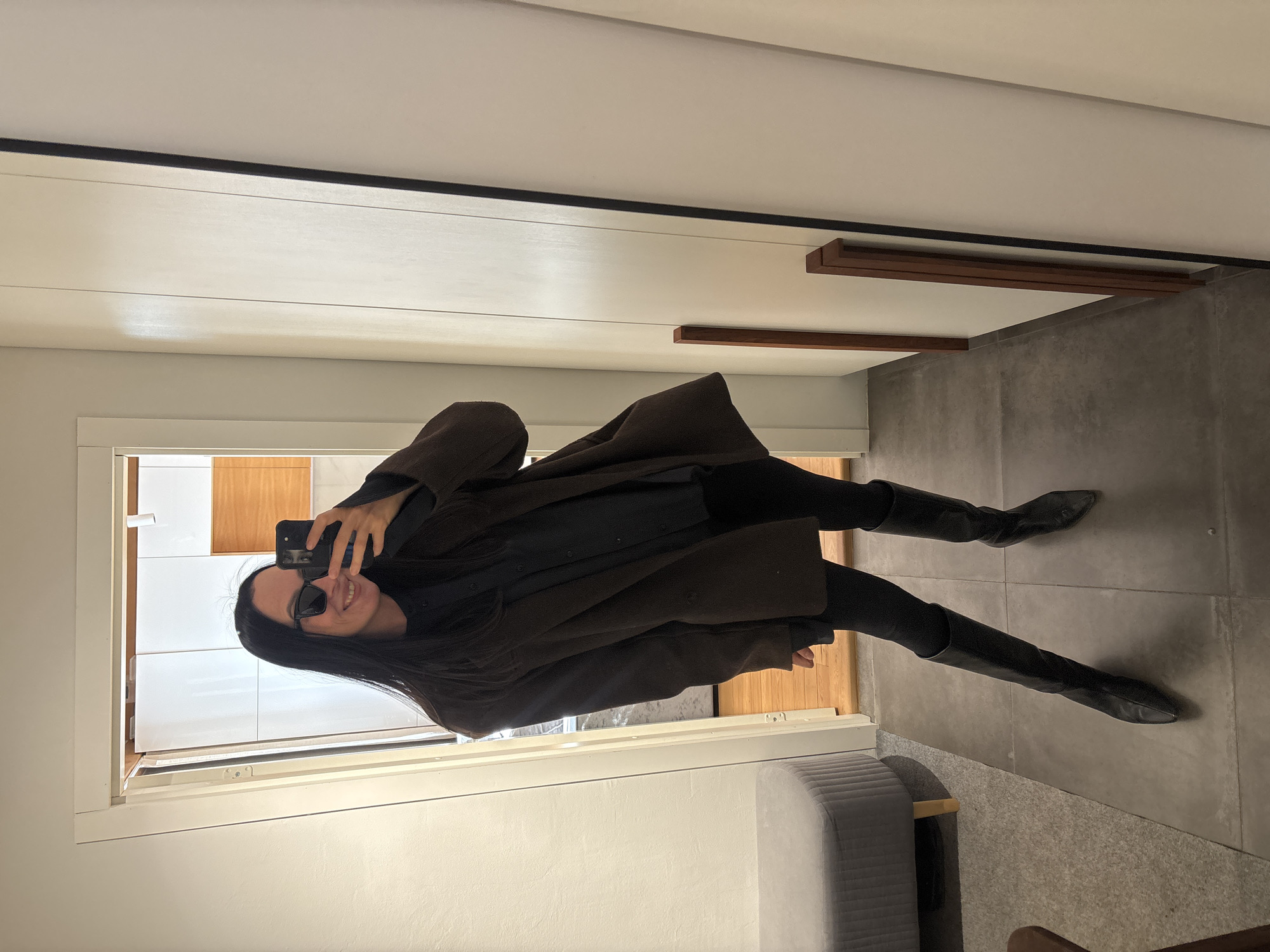 A woman is taking a selfie in a mirror. She is wearing black leggings, black knee-high boots, a black blouse, and a long brown coat. She is wearing sunglasses and smiling. The background includes a tiled floor, a gray bench, a white wall, and a glimpse into another room with white cabinets and wooden accents. There is no specific evidence in the image to place it in the city of Chaikovsky, Russia.
