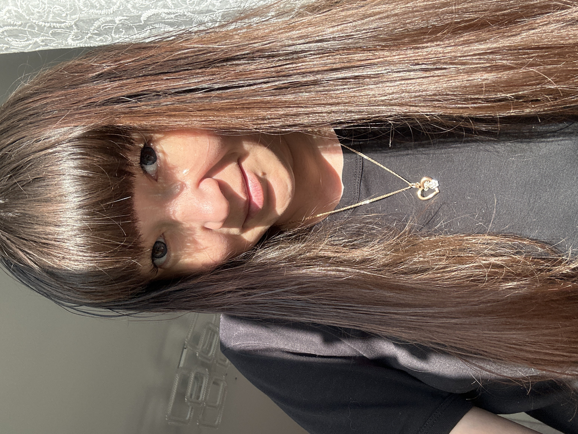 In an indoor setting, possibly a room in Chaikovsky, Russia, a woman with long, dark brown hair and bangs is taking a selfie. Sunlight casts shadows across her face, highlighting her features, including her dark eyes, nose, and lips, and her skin appears slightly shiny. She is wearing a black t-shirt and a gold necklace with a heart-shaped pendant adorned with a small gem. Behind her, a white lace curtain is visible, partially illuminated by the sunlight, and a patterned ceiling with square tiles can be seen.
