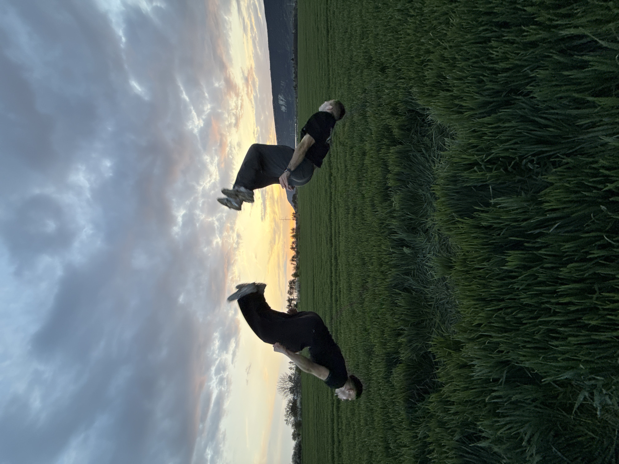 The image depicts two men in an open, grassy field at what appears to be dusk or dawn. The image is oriented with the horizon line running vertically. One man is in the middle of a backflip, with his body arched and his head nearing the ground. He is wearing a dark t-shirt and dark pants. The other man is in a standing position, slightly bent at the waist, with his feet off the ground. He is also wearing a dark t-shirt and dark pants. A mountain ridge is visible on the horizon, adjacent to a field. The sky is filled with clouds with colors ranging from gray to orange.
There is no indication in the image of the location being Shumen, Bulgaria.