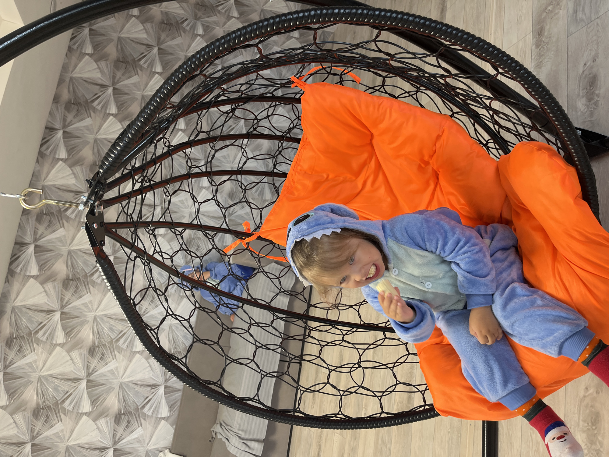 A young child in a blue shark pajama costume sits in a hanging egg chair with an orange cushion. The chair is suspended from the ceiling. The child is holding a small, yellow object. The room has a patterned wallpaper and light-colored wood flooring. The location is Chaikovsky, Russia.
