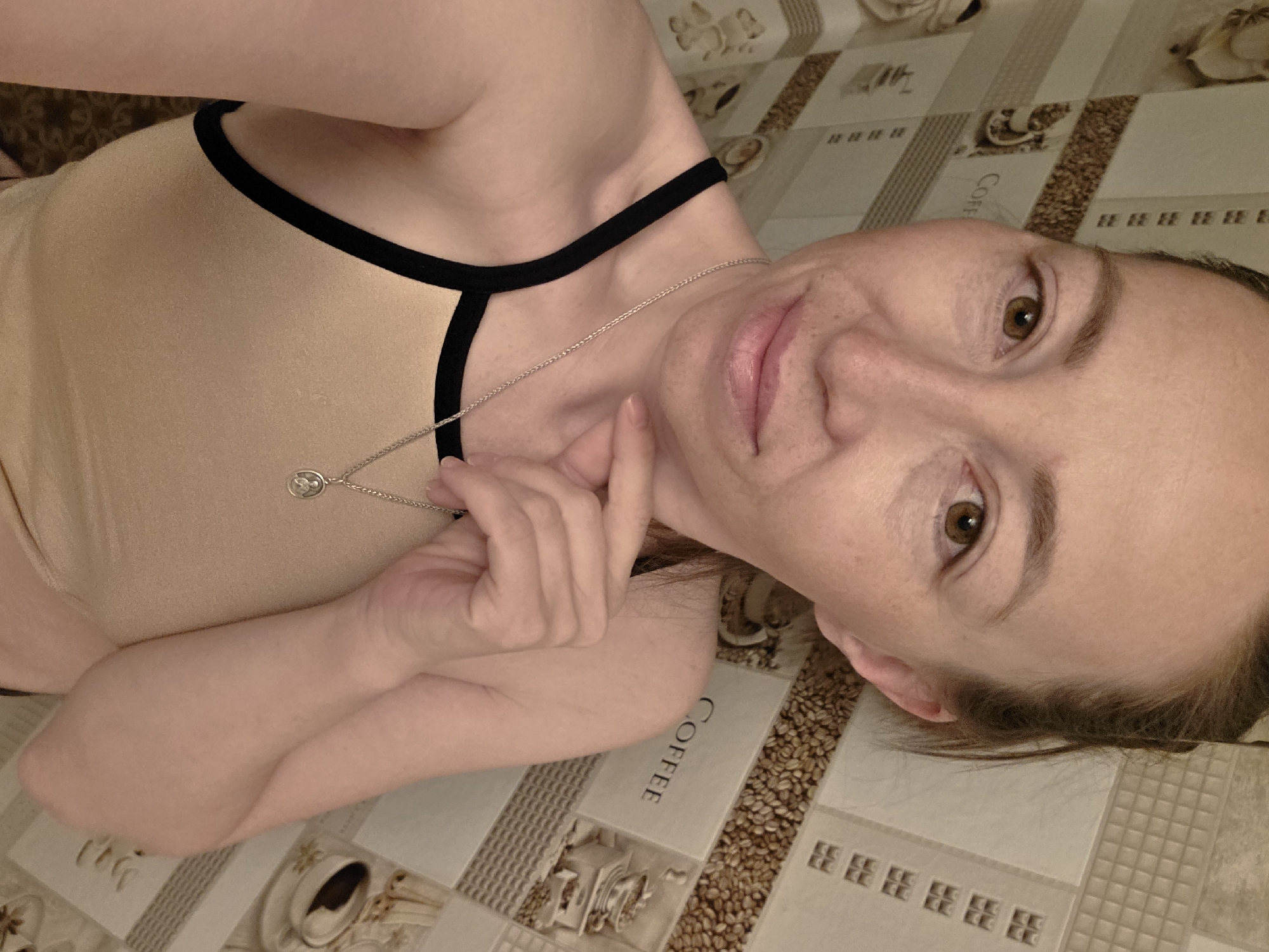 A woman lies on a surface patterned with images of coffee cups, beans, and the word "COFFEE". She wears a light-colored top with black trim and a silver necklace. Her right hand rests near her neck. She is facing the camera with a slight smile. The location is suggested as Chaikovsky, Russia.