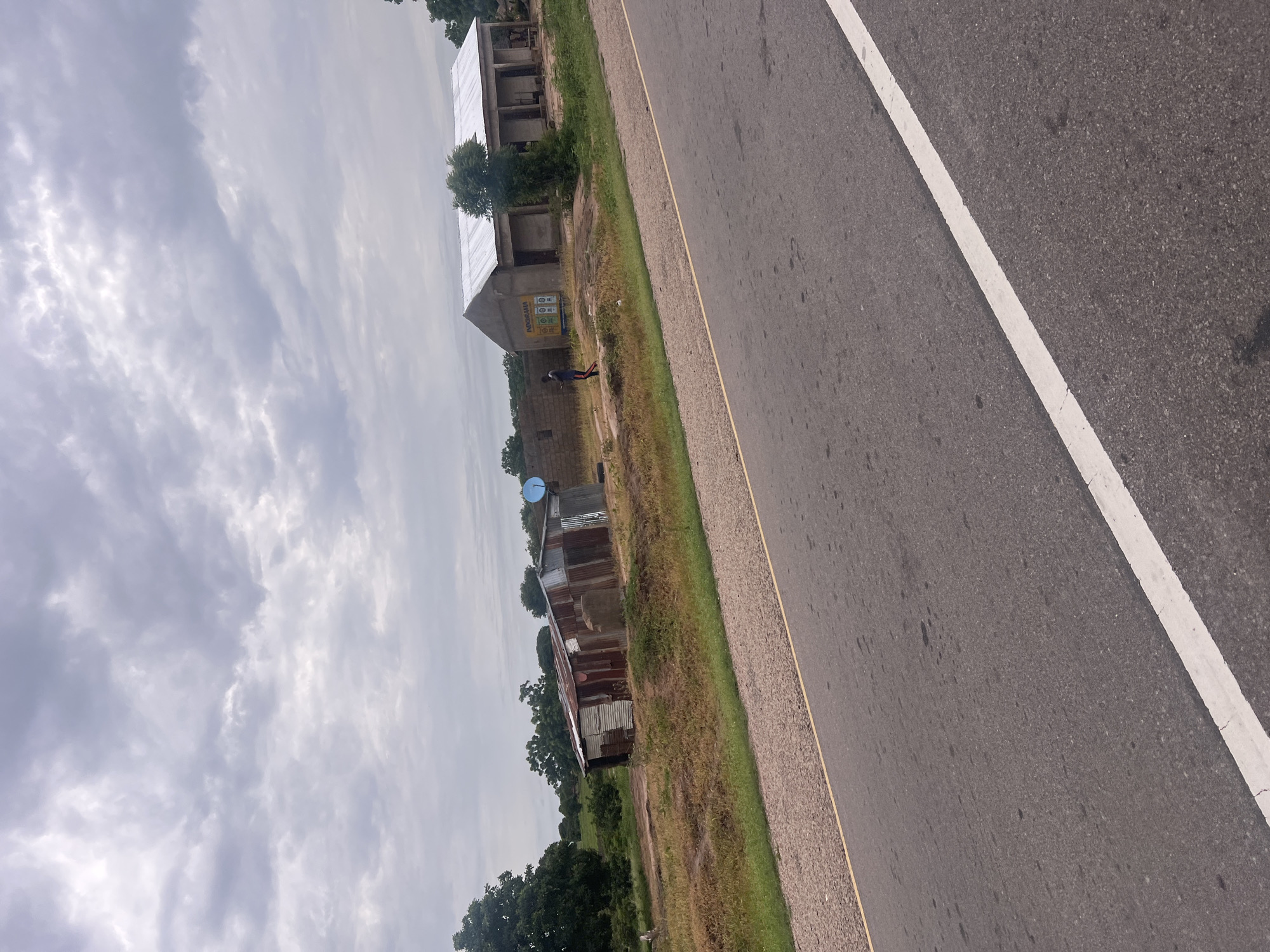 The image shows a roadside scene in Minda, Nigeria, under an overcast sky. The perspective is from a slightly elevated position, looking down at an angle. On the left side of the image, there is a dirt road or path running parallel to the paved road. Along the dirt road, several buildings are visible. The most prominent is a partially constructed building with a corrugated metal roof. Next to it are smaller, older structures, some appearing to be made of corrugated metal with rusted surfaces. One of these has a blue satellite dish on its roof. A single person, seen from behind, is standing near the partially constructed building. The person is wearing a blue shirt and dark pants. Behind the buildings, lush green trees are scattered, suggesting a rural or semi-rural environment.
The paved road occupies the right side of the image and has a white painted line marking its edge. The road surface is dark asphalt, with some scattered debris or stains. A narrow strip of gravel and grass separates the paved road from the dirt path. The weather appears cloudy and possibly humid, as indicated by the dull lighting and the type of clouds. The time of day is difficult to determine precisely, but the diffused light suggests it is not direct sunlight. The overall impression is of a developing area, with both new construction and older, more weathered structures present. There is a visible sign on the side of one of the buildings that appears to have text and logos, including what looks like "Nokia" and other circular emblems.