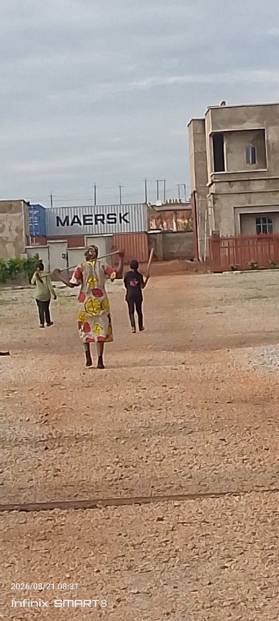 In Kakau, Nigeria, three individuals are observed in an outdoor setting. Two individuals are walking away from the camera, while one individual, positioned centrally, faces the camera. The central individual wears a brightly colored dress with a pattern of red, yellow, and green circles on a white background. This person holds two long, slender objects, possibly sticks, horizontally, one above the other, in front of their body, with arms extended upwards. To the left of the central figure, another individual walks away, wearing a light green long-sleeved shirt and dark trousers, also carrying a similar object horizontally over their left shoulder. To the right of the central figure, a third individual walks away, wearing a dark shirt with a graphic design and dark trousers, holding a similar object horizontally in their right hand.
In the background, shipping containers are visible, one prominently displaying the word "MAERSK" in large black letters. Behind the containers, a building with a light-colored facade and dark windows is partially visible, suggesting an industrial or commercial area. The ground surface appears to be a mix of gravel and dirt. The sky is overcast with clouds. A timestamp in the lower left corner reads "2026/08/21 08:31" and "Infinix SMART 8".
