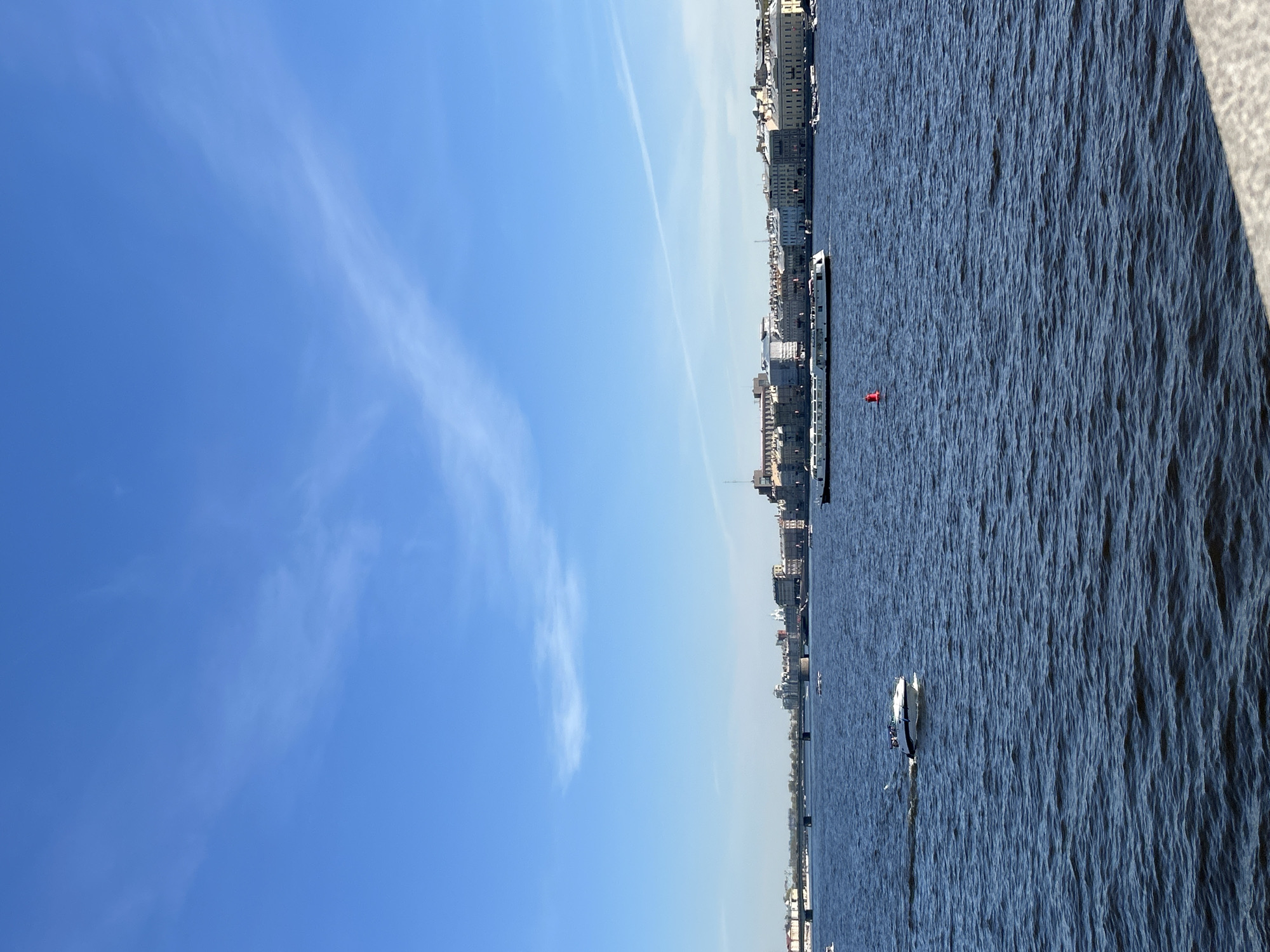 The image shows a view of the Neva River in Saint Petersburg, Russia. The sky is blue with wispy clouds. The buildings of the city line the riverbank. A barge is seen on the river, along with other smaller boats. The river's surface is rippled by the wind. A concrete barrier is seen in the top-right corner of the image.