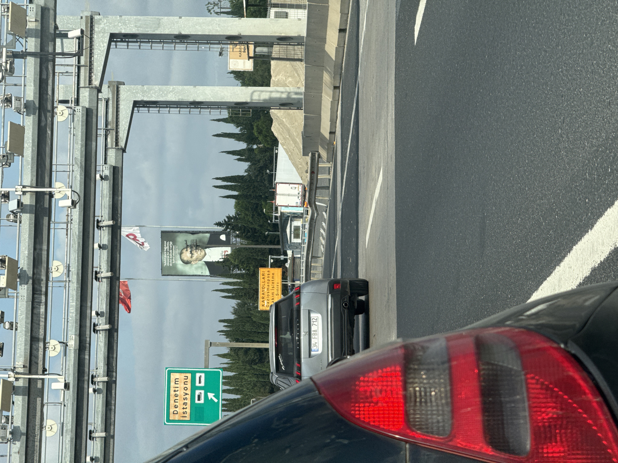 An outdoor, ground-level shot captures a highway scene with a toll booth structure overhead. In the foreground, the red taillight and rear quarter panel of a dark-colored vehicle are visible, along with a portion of the asphalt road and a white lane marker. Further ahead, a silver Peugeot SUV with license plate "34 FBA 712" is in motion. To the left of the SUV, a green road sign indicates "Denetim İstasyonu" (Inspection Station) with icons for trucks and a directional arrow pointing right. To the right of the SUV, a yellow sign reads "KARAYOLLARI Gaziosmanpaşa Akım İşletme Sınırı" (Highways Gaziosmanpaşa Flow Operation Limit). A large billboard featuring a portrait of Mustafa Kemal Atatürk is positioned behind the SUV, with Turkish flags visible in the background. The environment includes a line of tall, conical cypress trees and a cloudy sky. The toll booth structure itself is a metal framework with visible cameras and lights.