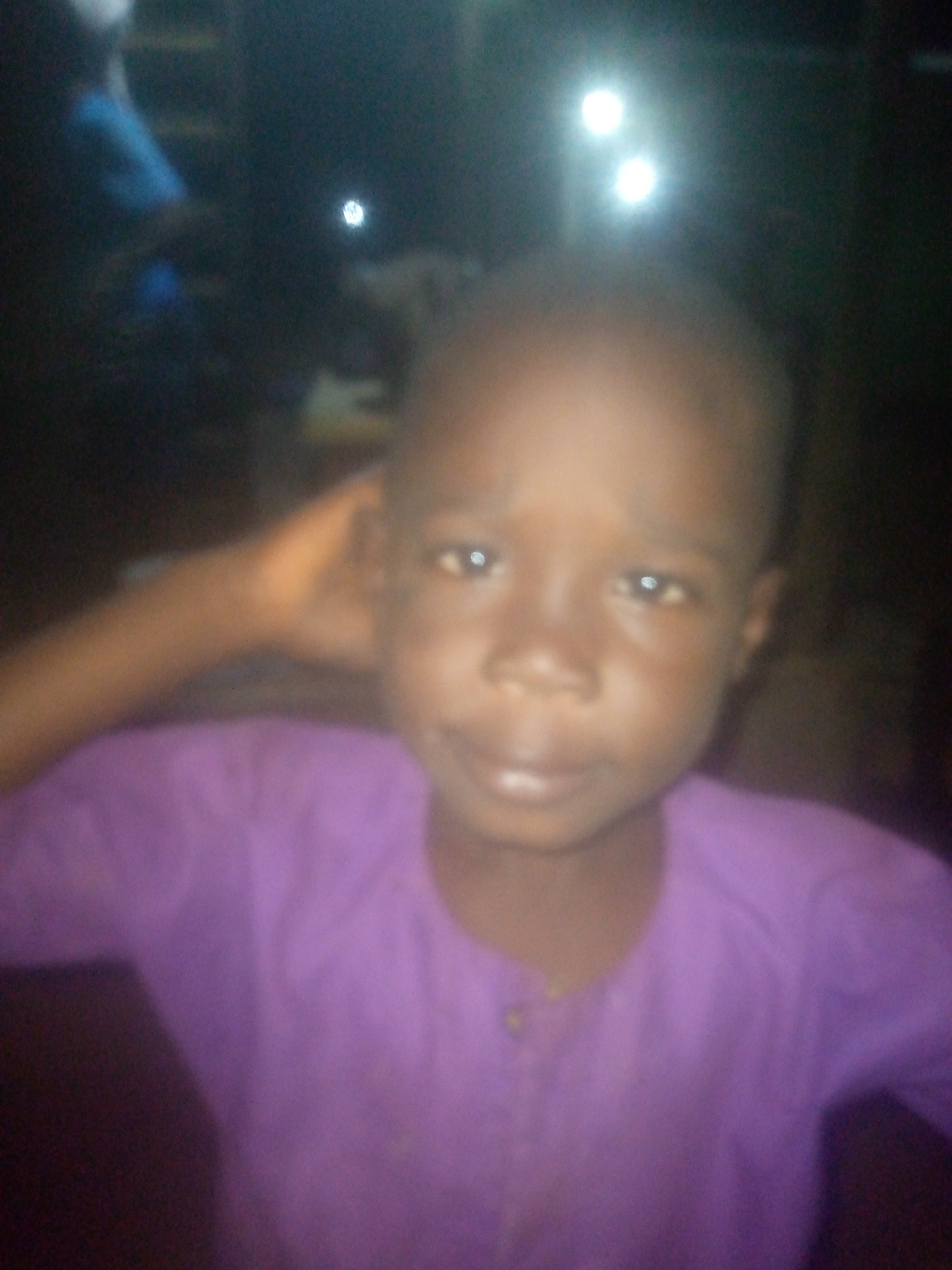 A portrait-style image captures a young child, likely male, with dark skin, looking directly at the camera. The child is wearing a purple, long-sleeved garment, possibly a shirt or tunic, with a simple round neckline and what appear to be small buttons or clasps at the front. A subtle smile plays on the child's lips, and the eyes show glints of light from reflections. One arm is slightly raised, with the hand positioned near the side of the head on the right side of the frame (the child's left).
The background is predominantly dark and very blurry, suggesting a low-light environment, possibly night-time or an indoor setting. Several bright, out-of-focus light sources appear as glowing white orbs in the upper and right portions of the background. A faint hint of blue is visible in the upper left background, possibly indicating another person or object obscured by darkness and blur. The overall image has a soft focus, with the child's face being the clearest element. There is no visible text.