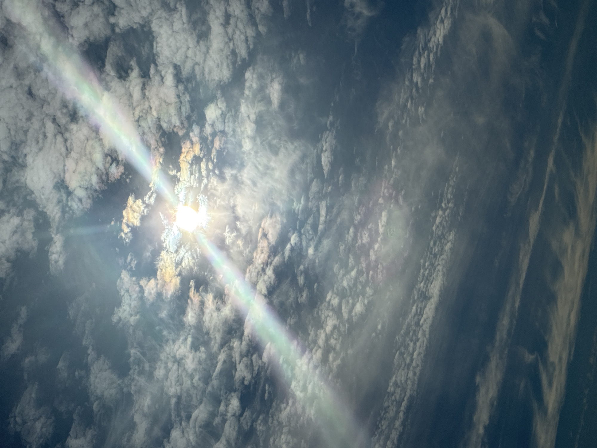 The image depicts a sky filled with a variety of clouds, ranging from dense, puffy formations to thin, wispy streaks. The sun is visible, peeking through the clouds and casting a brilliant beam of light that splits into a rainbow-like spectrum, suggesting a possible atmospheric phenomenon like a sun dog or iridescence. The overall lighting suggests it is daytime. The scene is devoid of any human presence or urban elements, focusing solely on the natural beauty of the sky. The location is unknown.