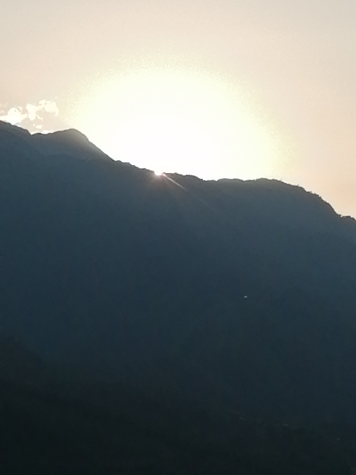 The image presents a serene mountainous landscape at either sunrise or sunset, with the location specified as Sa Pa Ward, Vietnam. A vast, dark mountain range occupies the majority of the frame, appearing in silhouette against the bright sky. The sun, an intensely bright and almost blinding light source, is positioned directly behind the jagged mountain ridge. Its brilliant light emanates upwards, casting a warm, hazy glow across the sky, which transitions from bright white near the sun to a softer, lighter tone higher up. Distinct sunrays are visible peeking over the mountain peaks, particularly towards the right side of the image. On the far left, a small, wispy cloud formation can be seen just above one of the mountain peaks. The overall impression is one of natural beauty and tranquility, characteristic of the mountainous terrain found in Sa Pa. There are no visible people, animals, or text in the image.