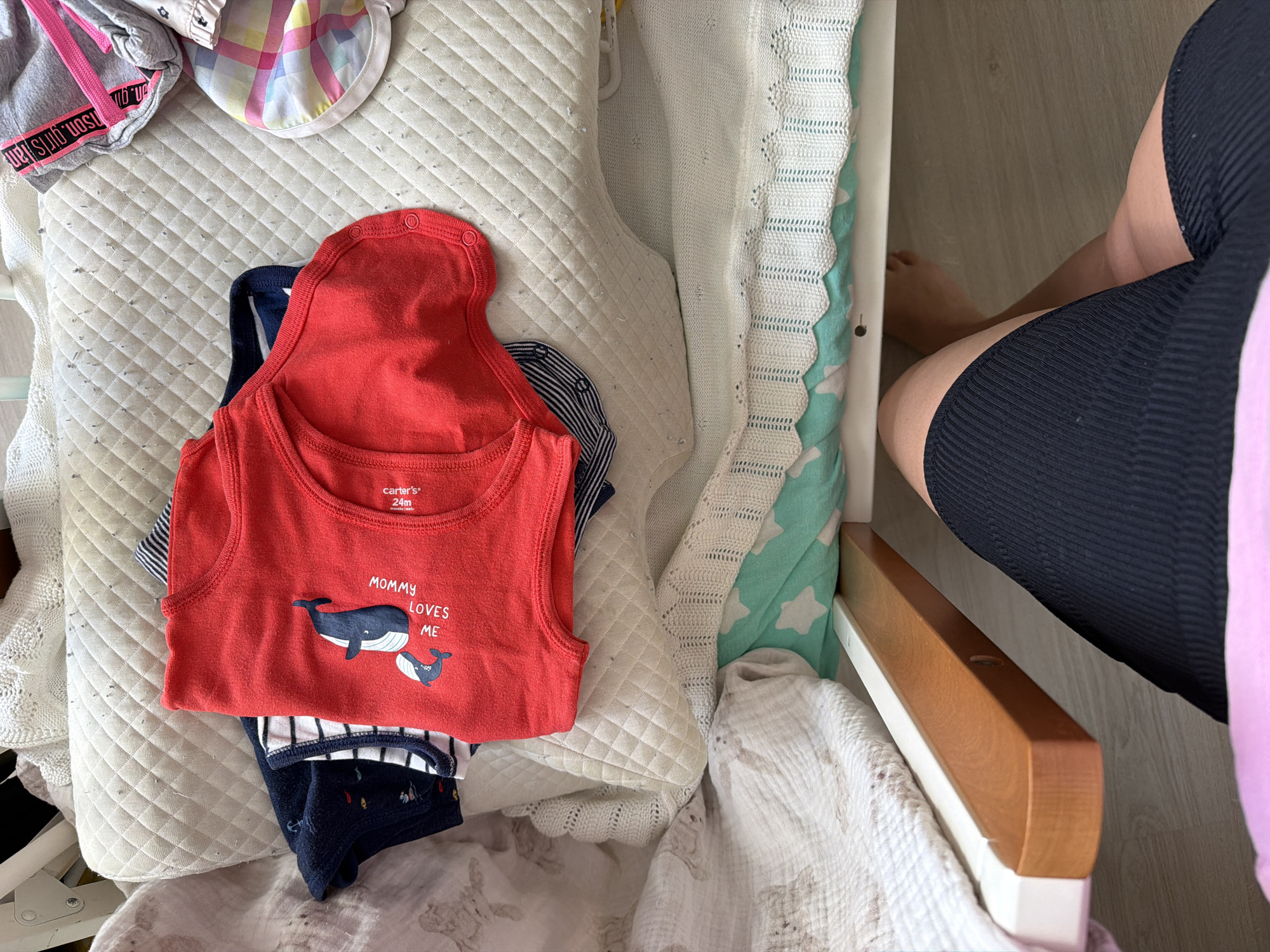 An overhead indoor shot captures a domestic scene, likely in a nursery or bedroom in Sochi, Russia. The central focus is a white, quilted padded surface, possibly a changing pad or part of a crib mattress, on which several baby clothes are laid out.
Dominating the center is a red sleeveless bodysuit or tank top, featuring a graphic of a large and small whale and the text "MOMMY LOVES ME". The label "carter's 24m" is visible on the neckline. Beneath the red garment, a navy and white striped bodysuit is partially visible, along with another dark blue fabric patterned with small fish or marine life. To the top left, a grey garment with a pink drawstring and partially visible text (reading "son. girls han" and "n. girls" in reverse) is folded. Next to it, a colorful, checkered baby cap or hat is placed.
To the right of the padded surface, a person's lower body is visible, wearing dark, ribbed athletic shorts or leggings. Their bare feet are visible on a light wooden floor beneath a wooden crib rail. A white crocheted blanket and a light green blanket with white star patterns are also visible draped over parts of the crib structure. The lighting suggests it is daytime. The overall impression is a casual, everyday moment of childcare.