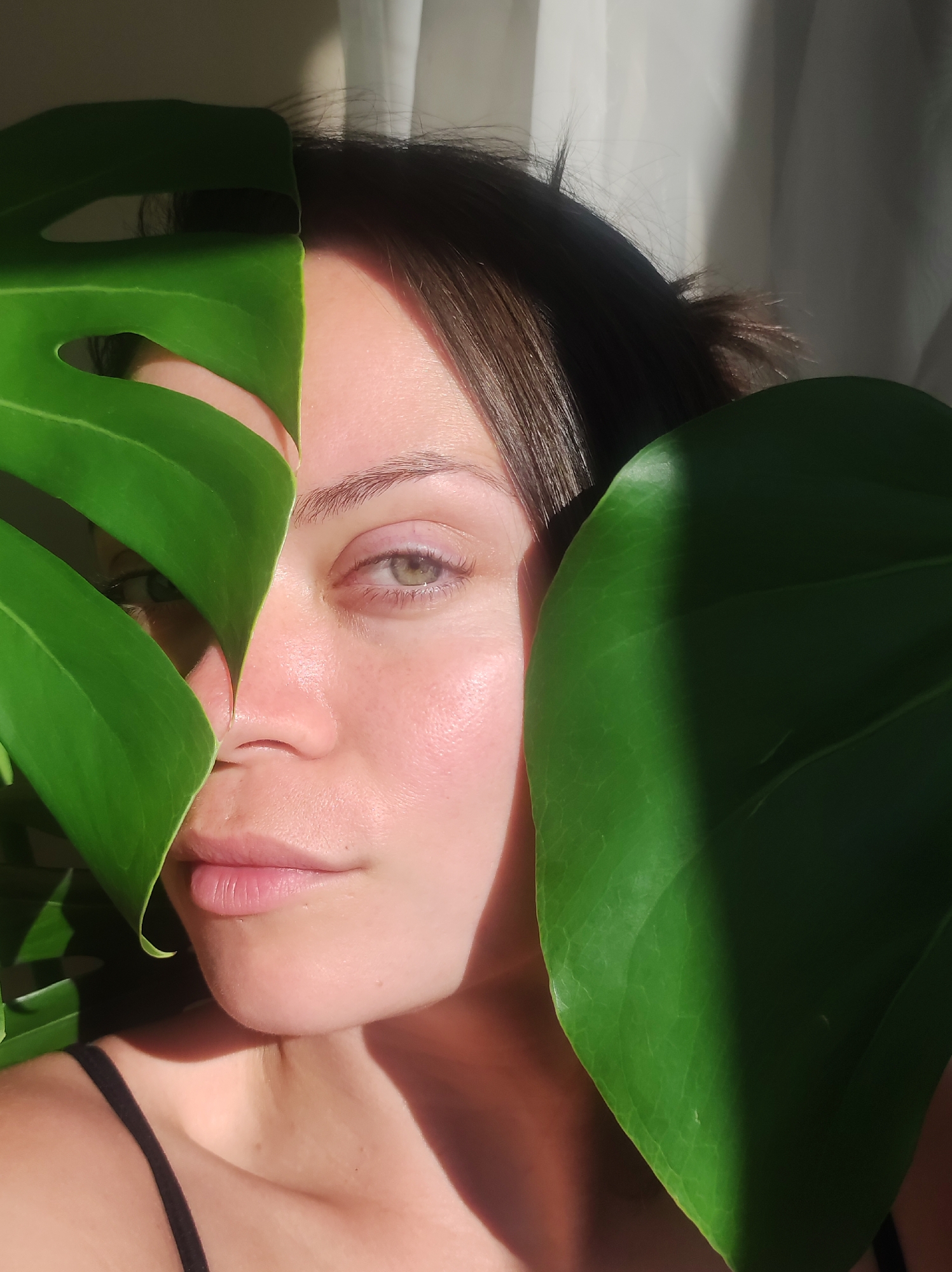 The image shows a close-up of a woman's face, partially obscured by large green leaves. Her left eye and part of her nose and mouth are visible. She has light-colored skin with a rosy blush and light brown hair. The background consists of a softly lit curtain. The lighting suggests a natural light source, possibly from a window. There are no visible clues in the image to suggest a specific location within Chaikovsky, Russia.