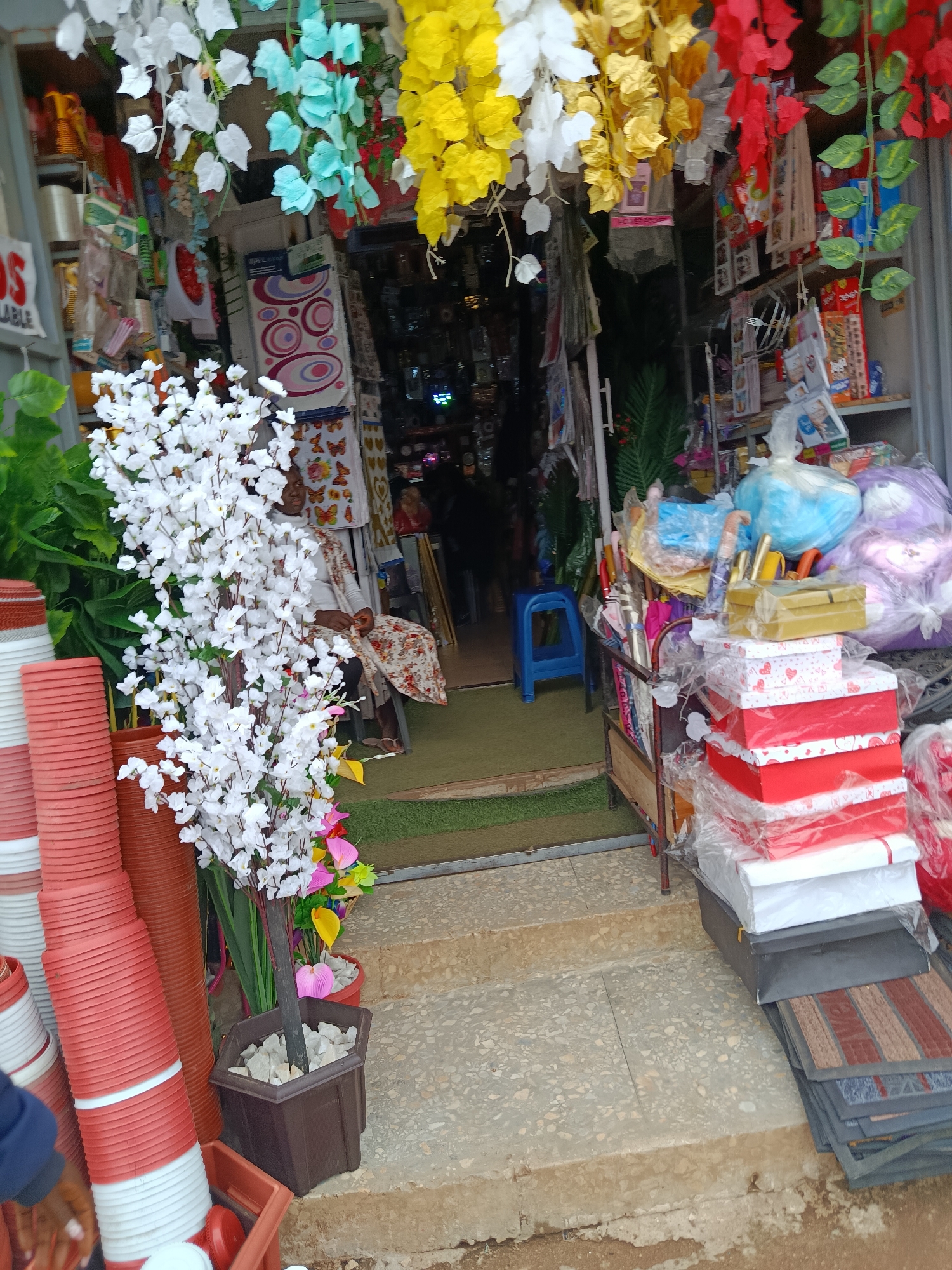 The image captures a storefront in Jos, Nigeria, displaying a variety of items. In the foreground, a stack of red and white plastic pots is visible on the left, next to a decorative white flowering plant in a brown pot. To the right, a collection of stacked gift boxes, predominantly red and white with heart designs, are piled high, some still wrapped in plastic. Several umbrellas are leaning against a metal stand behind the gift boxes. In the background, the interior of the shop reveals shelves filled with colorful artificial flowers in yellow, blue, and red. A person is seated inside the shop on the right, partially obscured by the decorative plants. The ground appears to be a concrete surface with some rough-hewn steps leading into the store. The overall scene suggests a retail environment specializing in decorative items and household goods.