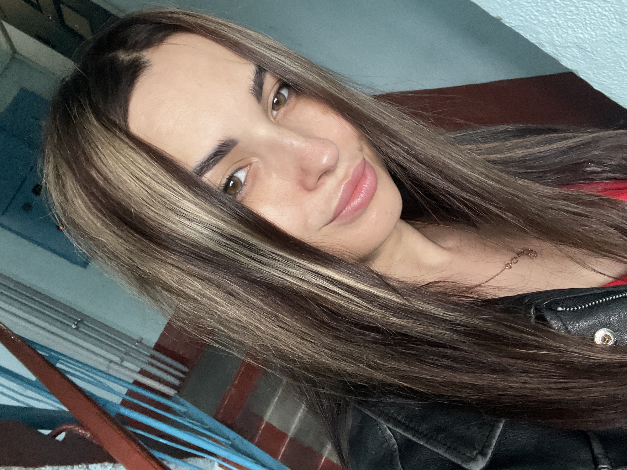 A young adult female is depicted in a close-up, chest-up portrait. She has long, dark brown hair with distinct blonde/caramel highlights framing her face and running through the length. Her eyes are brown, and her eyebrows are dark and well-defined. She exhibits a subtle smile with full, pink lips. Her skin has a light-medium tone, showing minor texture and slight discoloration in areas. She is wearing a black leather jacket over a visible red garment. A delicate gold-colored chain necklace with small, linked charms is visible around her neck. She gazes directly at the camera.
The background suggests an indoor location, specifically a stairwell or hallway. To the left, a section of a reddish-brown stair railing is visible, behind which are vertical blue metal balusters and hints of steps or wall elements. The wall on the right side of the frame is a faded light blue or teal color, with a darker, reddish-brown vertical section present behind her right shoulder and hair. The setting appears to be within a building, possibly a residential or institutional structure, in the city of Unknown, Unknown.