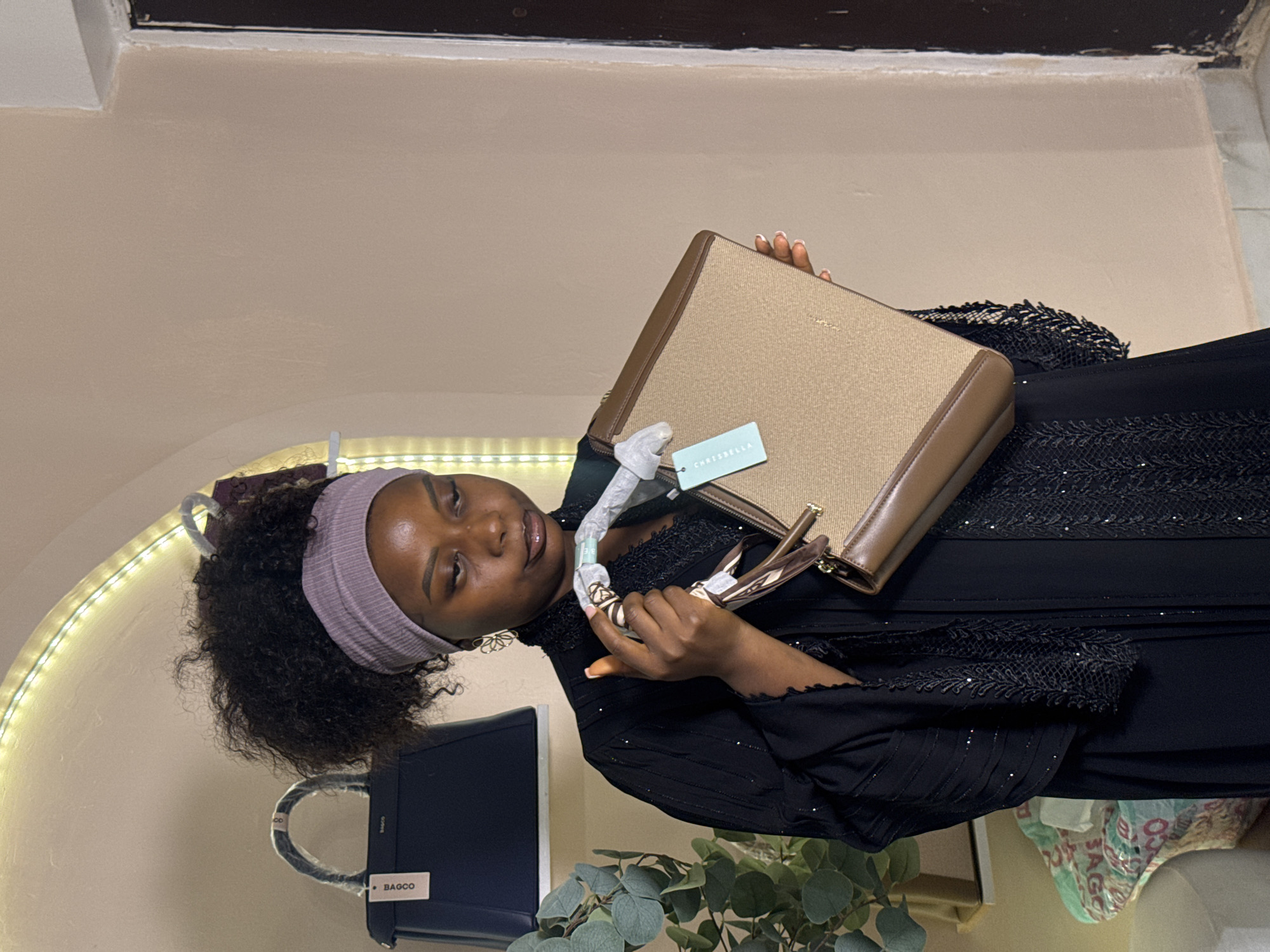 In an indoor setting, a young Black woman is captured in a portrait. She is wearing a light purple headband and a black, sequined dress. The woman is holding a light brown, rectangular handbag with a metallic sheen. She appears to be examining the handbag, perhaps showing it off or considering a purchase.
In the background, a dark blue handbag with the label "BAGCO" is visible. Additionally, a string of warm-toned fairy lights arches across the upper left portion of the frame, suggesting a decorative element or ambiance. Some greenery, likely artificial plants, is present in the lower right corner of the image. The overall impression is one of casual display or review of accessories in a retail or personal collection environment. The lighting is soft and ambient, with no clear indication of the time of day or weather conditions. The scene is indoors, with plain, light-colored walls forming the backdrop.
Visible text includes "CHRISBELLA" on a tag attached to the handbag the woman is holding, and "BAGCO" on the other handbag.