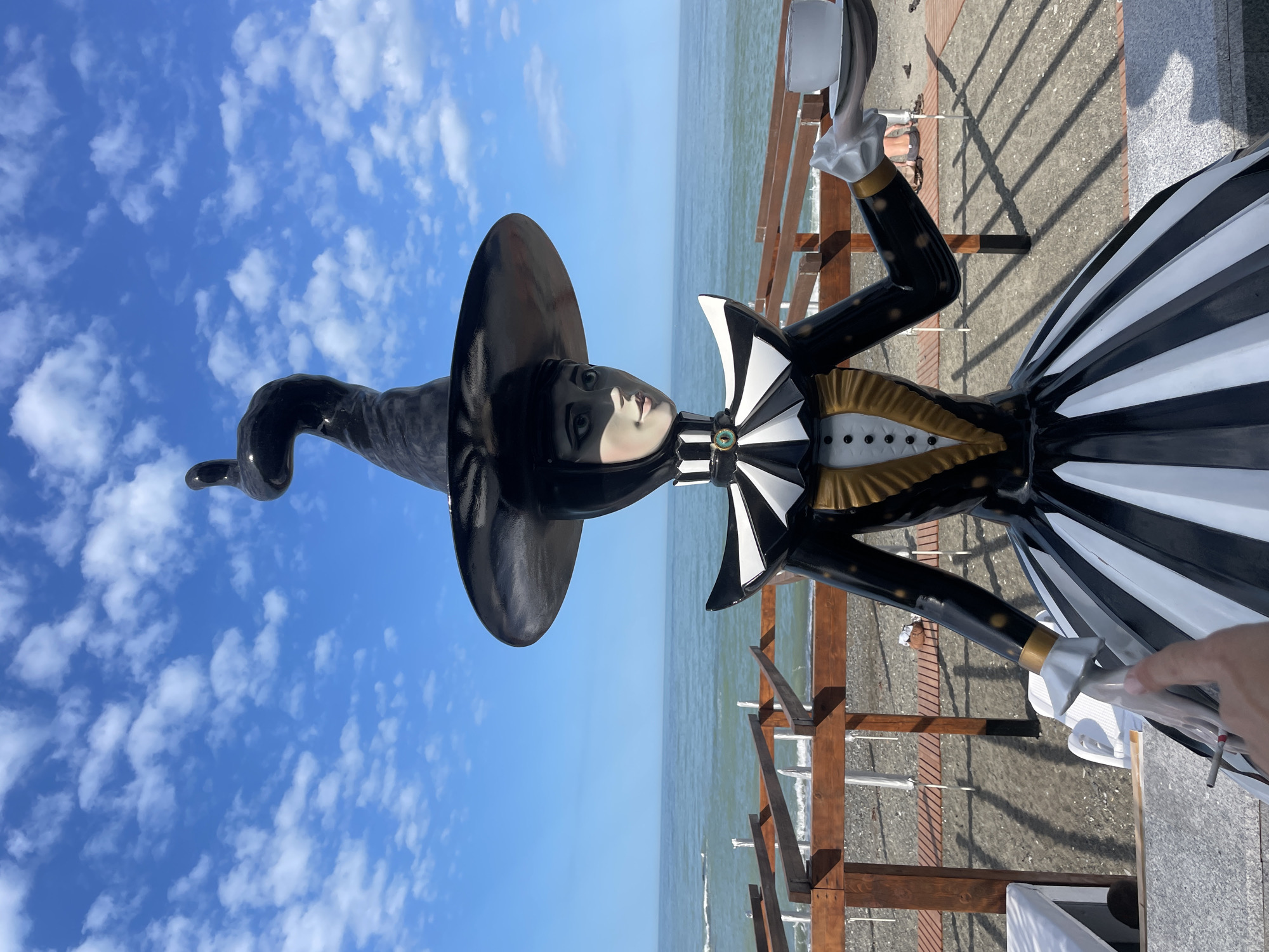 The image shows a statue of a woman wearing a black and white harlequin-style dress, a large, black witch's hat, and holding a white object, possibly a cup or a small container. The statue is positioned outdoors, with a view of the sea and sky in the background. The sky is blue with scattered white clouds. In the background, there's a wooden structure and a pebbled beach. The statue is located in Sochi, Russia.