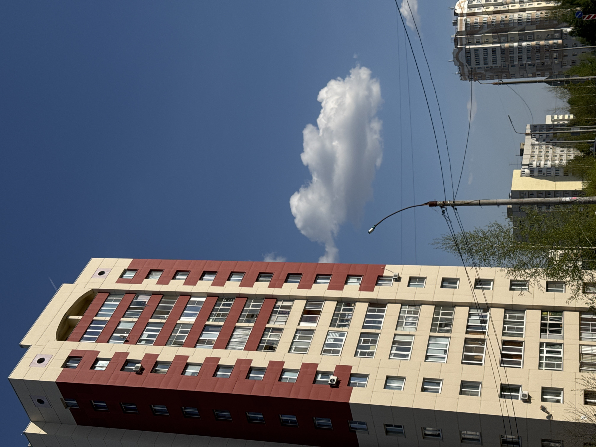 The image shows a tall, modern building in Kazan, Russia. The building has a cream and red facade with multiple rows of windows. The sky is blue with a single white cloud visible. In the background, there are more buildings, power lines, and trees. A streetlight pole stands near the building.