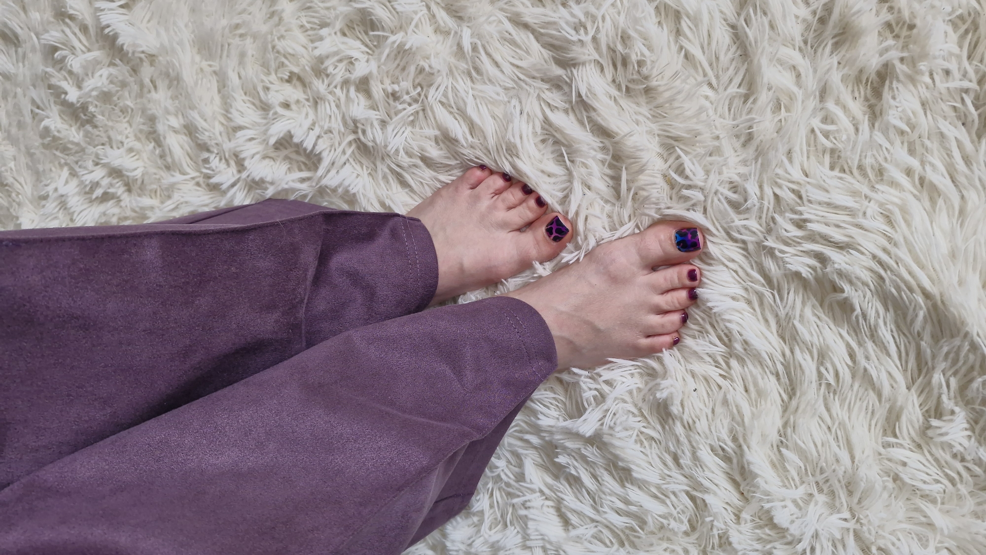 A top-down indoor shot shows a person's legs and feet resting on a fluffy white rug. The person is wearing purple pants. The feet have polished toenails, with most of them in a dark purple color, and one big toenail on each foot painted with a colorful pattern of blue, purple, and pink. The rug has a shaggy texture with visible strands of white. No other objects or surroundings are visible, making it impossible to determine the exact location in Otchor, Russia.