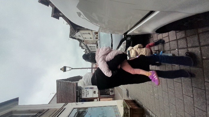 The scene depicts an urban environment on a cloudy day in Schiltigheim, France. On a sidewalk paved with rectangular grey blocks, an adult figure, seen from behind, wears a black coat and a white knit beanie with a pompom. This adult carries a small child, also seen from behind, dressed in a pink puffy jacket, light pink leggings, and purple Adidas sneakers. The child wears a dark knit beanie with a pompom. To the right of the adult, a second small child, seen from behind, wears a tan puffy jacket, red patterned leggings, and black boots, and is seated on a small blue and red ride-on toy.
In the background, a dark-colored car is partially visible on the street behind the individuals, and a large white van occupies the right foreground, obscuring part of the street. A traditional black streetlamp with a brass-colored cap stands in the middle ground. Buildings line the street, including a structure on the left with a display window containing text, and buildings with brown-tiled roofs extending into the distance under an overcast sky. A wooden planter box is situated on the left side of the sidewalk.