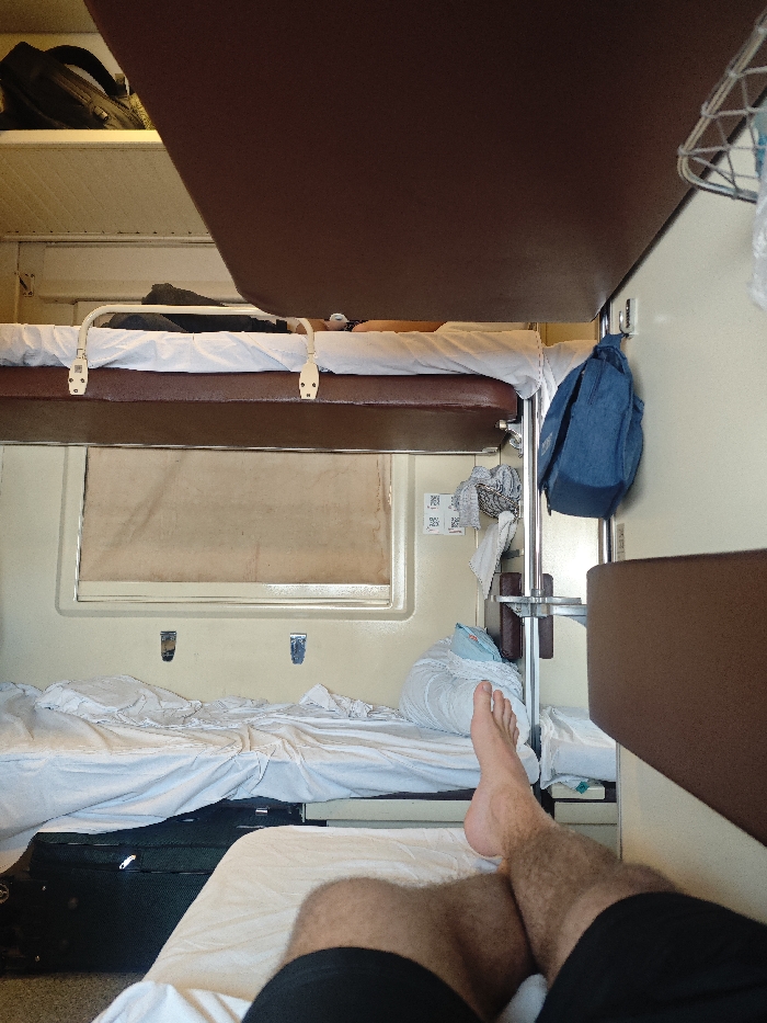 The image depicts the interior of a passenger train sleeping compartment, specifically located within or passing through the city of Druzhinino, Russia. The perspective is from a lower bunk, looking towards the foot of the compartment. In the immediate foreground, the bare feet and lower legs (clad in black shorts) of an individual are visible, resting on a white-linened bunk.
Above this, another lower bunk is prepared with white sheets, a pillow, and a folded blanket. A black hard-shell suitcase with wheels is stowed underneath this bunk. Centered on the wall beyond the lower bunk is a window with a light-colored, opaque shade fully drawn. To the right of the window, a light-colored wall features two small metal hooks. Further right, a vertical metal pole supports a brown shelf, a wire basket containing items (possibly laundry or personal effects), and a blue backpack hanging from a hook. Two small square white stickers with black markings are affixed to the wall above the shelf.
An upper bunk, also made with white linen and a headrest, is visible above the lower bunk. A second individual is lying prone on this upper bunk, with their head and arm partially visible. Above the upper bunk, a brown overhead shelf holds a black backpack. A large, dark brown panel extends from the ceiling, partially obscuring the upper section of the image frame. The compartment walls are a light beige or cream color.
