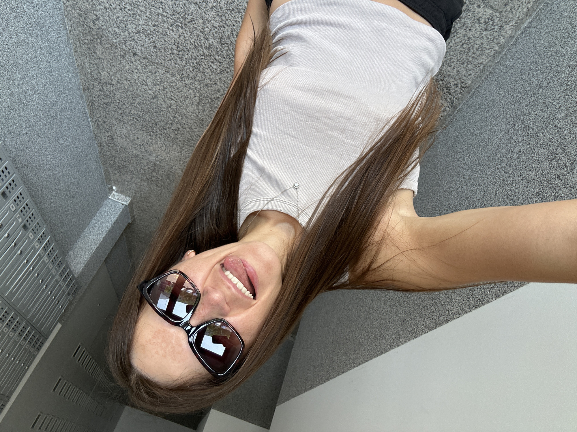 This is a selfie taken from a low angle by a young woman with long, dark brown hair. She is wearing oversized, dark-rimmed sunglasses and a light-colored, ribbed tank top. Her mouth is open, and her tongue is slightly sticking out, suggesting a playful or energetic mood. She has a delicate silver necklace with a small charm visible against her top.
The background appears to be a staircase with textured grey surfaces. To the left, a row of lockers with many small square openings is visible. The lighting suggests it might be daytime, possibly indoors or in a shaded outdoor area. The overall impression is casual and candid. There is no visible text in the image.