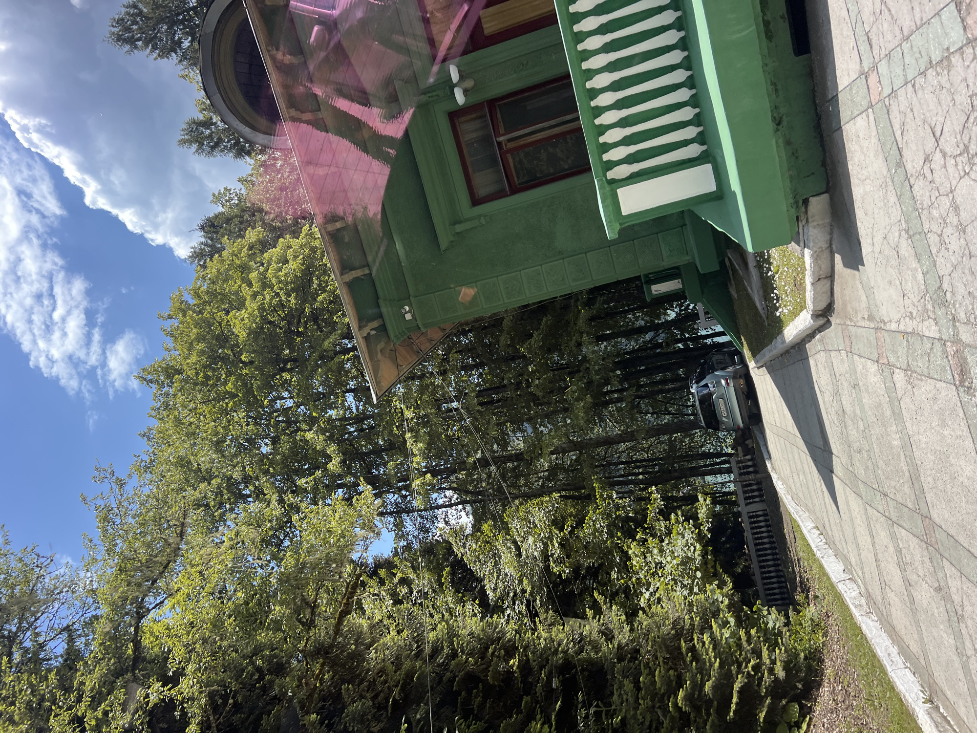 This media file depicts an outdoor scene with a focus on a green, two-story house. The house features a red-trimmed window and a distinctive white-railed porch or balcony. A pink-tinted, transparent canopy extends from the house, reflecting the sky and trees.
Surrounding the house is lush green foliage, including large trees with dense canopies and smaller bushes. The ground in front of the house appears to be a paved walkway with a pattern of grey and pinkish tiles. A small patch of grass with white flowers is visible along the edge of the walkway and house.
In the background, tall trees are visible, and a dark-colored SUV is parked on a road or driveway, partially obscured by the trees. The sky is blue with scattered white clouds, suggesting it is daytime, likely morning or afternoon, with fair weather. The overall atmosphere is peaceful and natural. There are no people visible in the image. The image is taken from a slightly elevated perspective, looking downwards and to the side.
There is no discernible text visible in the image. The scene appears to be set in a location with abundant vegetation, possibly a residential area or a park. The orientation of the image is rotated, so the house and walkway appear to be on a steep incline.