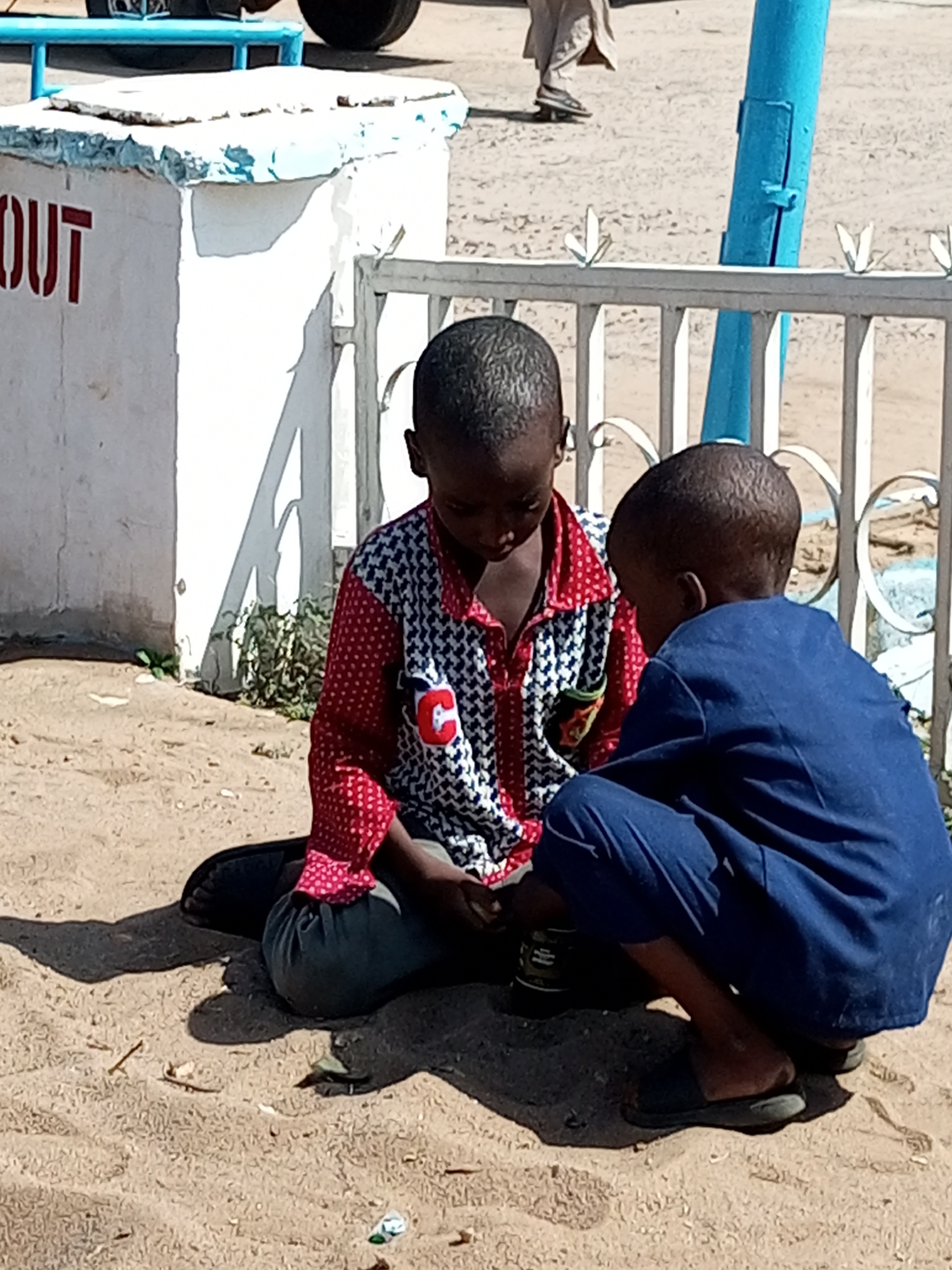In Garko, Nigeria, two young boys are seated on the sandy ground near a white, concrete structure with red lettering that reads "OUT." The boy on the left wears a red and white patterned shirt and dark pants. He is looking down, concentrating on something between his hands. The boy on the right is dressed in a blue outfit and is also looking down. They appear to be interacting with something on the ground in front of them. Behind the boys, a white metal fence with decorative elements separates them from a blue pole and a dirt surface. The environment suggests an outdoor, possibly public, area.