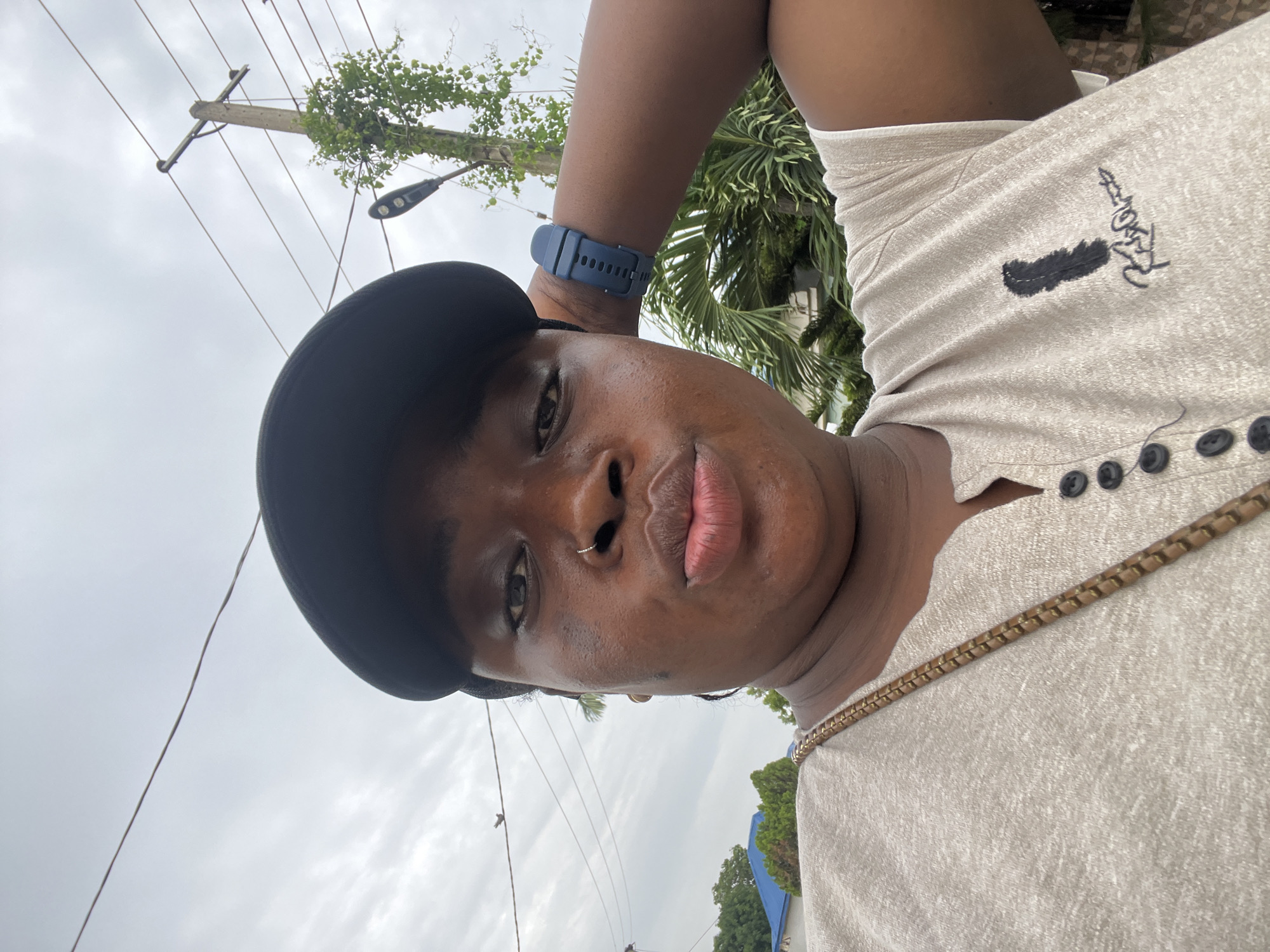 A person wearing a black baseball cap, a light-colored, button-down shirt with a gold chain, and a blue smart watch is shown in a low-angle, close-up shot. The person has a nose ring. In the background, there are utility poles with wires, green trees and vegetation, and a cloudy sky. This image appears to have been taken outdoors, potentially in Abuja, Nigeria.