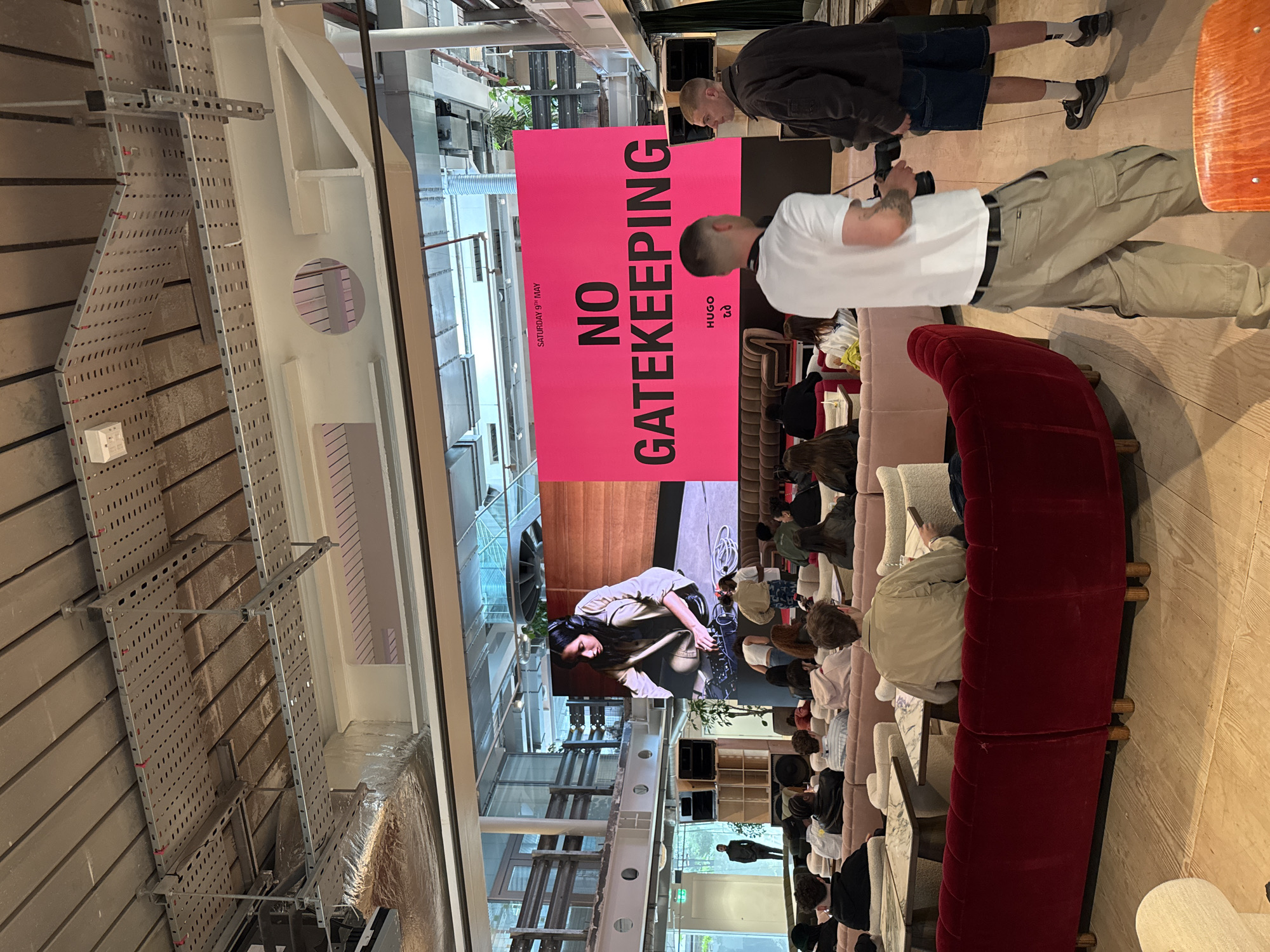 This indoor shot shows a crowd seated on various sofas and chairs, facing a large screen where a woman with headphones is featured. A pink banner reads "NO GATEKEEPING, SATURDAY 9TH MAY, HUGO e3." Two men stand to the right; one is taking a photograph. The event appears to be taking place in a building with an open, industrial ceiling, located in London, United Kingdom.