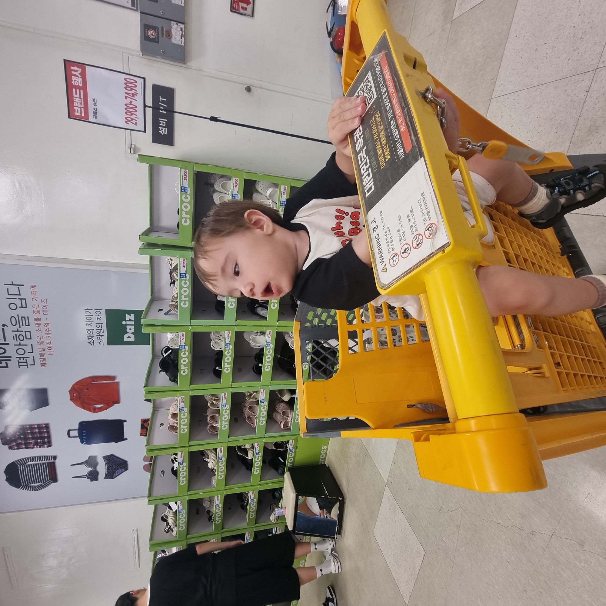 In a retail store in Wonju-si, South Korea, a young child sits in a yellow shopping cart. The child wears a black and white t-shirt and is facing to the right, with their mouth slightly open. The shopping cart has a yellow handle and a black interior grid. Attached to the side of the cart is a warning sign with Korean text and icons depicting children. The background shows rows of shelves filled with footwear, primarily branded with "crocs." Some of the footwear is displayed in green boxes. A large promotional poster hangs on the wall behind the shelves, featuring images of clothing and a suitcase, with Korean text. Another sign with Korean text is visible on the wall above the footwear shelves. A person's legs and feet, wearing white socks and black sneakers, are visible on the left side of the image, standing near the shelves. The floor is tiled with a light gray pattern.