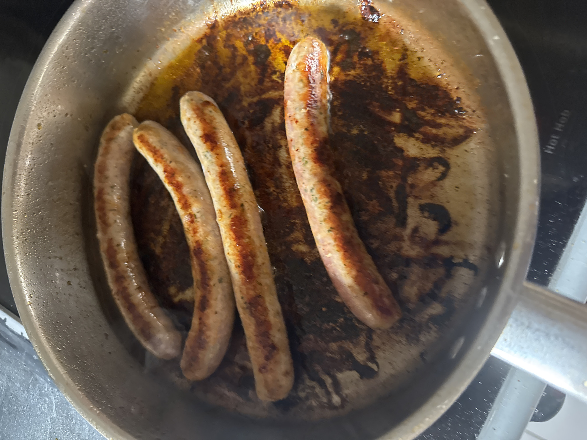 The image shows four sausages cooking in a frying pan. The pan appears to be stainless steel and sits on a stovetop. The sausages have a light brown, cooked exterior with some browned spots. The pan contains a significant amount of grease and some dark, burnt residue. The environment is a kitchen setting, though the exact location within Paris, France, is not discernible from the image.