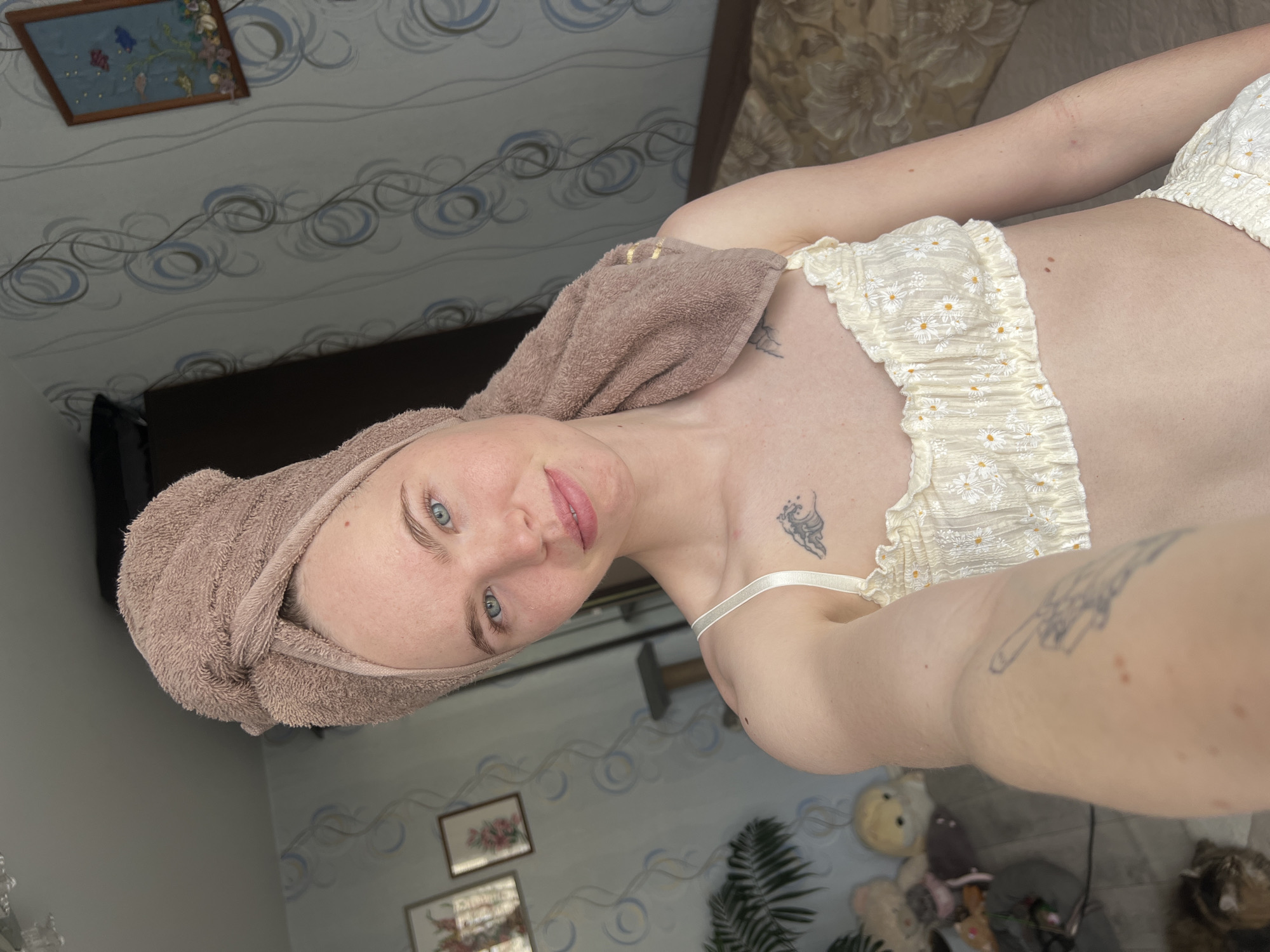 A selfie of a fair-skinned woman, possibly in Chaikovsky, Russia, is oriented vertically. She has a light brown towel wrapped around her head. She is wearing a white top with small yellow flower designs. The woman has blue eyes and light pink lips. Tattoos are visible on her chest and arms. The background includes patterned wallpaper with floral and swirl designs, a framed picture on the wall, a dark-colored television, a fern-like plant, and several stuffed animals.