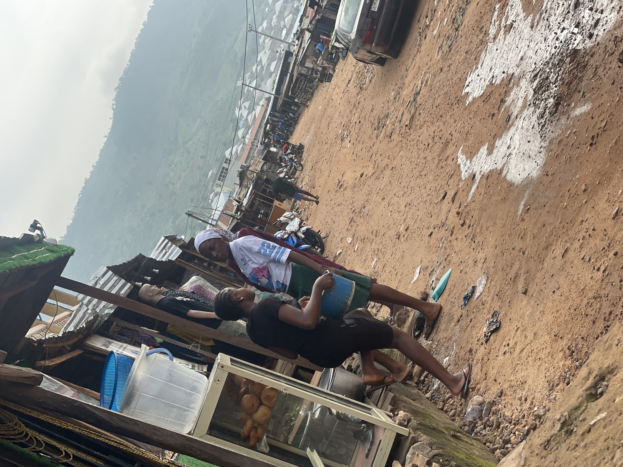 The image captures a bustling street scene in Lokoja, Nigeria, with a focus on a roadside market. In the foreground, two young women are interacting. One, wearing a black t-shirt and shorts, is bent over, holding a blue bucket. The other, in a white t-shirt with blue lettering and a green skirt, stands next to her. To the left, a glass display case filled with pastries or fried dough sits on a wooden structure. Beyond them, the street is lined with various market stalls constructed from corrugated metal and wood, some with awnings. Several motorcycles are parked in the area, and a few people are visible walking through the market. In the background, a car is parked on the right side of the dirt road. The scene is set against a backdrop of a hazy, mountainous landscape dotted with houses, indicating the elevated terrain of Lokoja. The ground is primarily red, dusty earth, with some scattered debris. The overall atmosphere suggests a vibrant, everyday commercial activity in this Nigerian city.