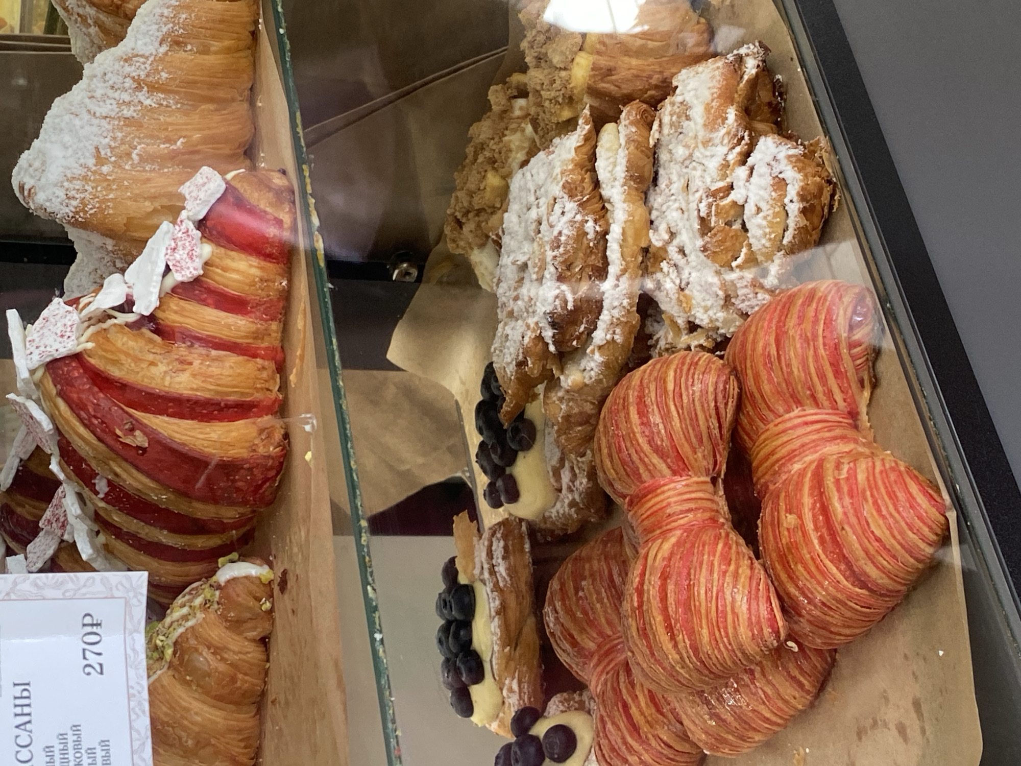 The image shows a display case filled with pastries, likely in a bakery or café. The pastries include croissants with red stripes, some topped with white decorative pieces, and others dusted with powdered sugar. Some croissants are filled with a cream and decorated with blueberries. A price tag is visible, indicating a price of 270 rubles. The location of the bakery or café is suggested to be in Votkinsk, Russia based on the image request.