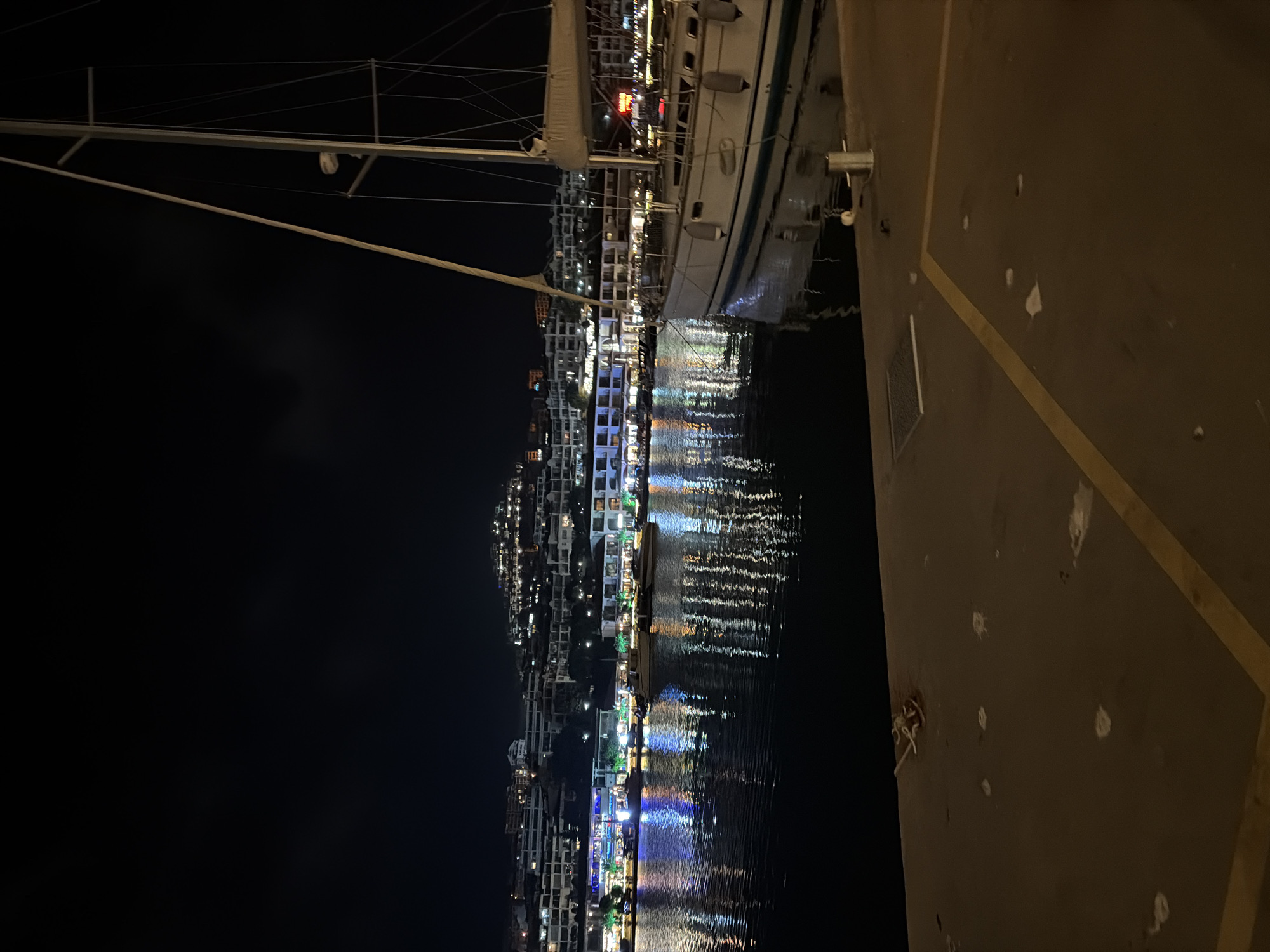 This nighttime image depicts a marina in Sveti Vlas, Bulgaria. A white sailboat with a blue stripe along its hull is moored on the right side of the frame, occupying the midground. Multiple dark fenders hang from its starboard side. Its mast and rigging extend vertically into the dark sky, featuring a furled sail on the boom and an electronic device mounted on the mast.
The foreground consists of a dark, paved dock surface. Yellow painted lines form an L-shape on the surface, and scattered white debris or marks are visible. A silver-colored bollard is present on the dock to the right, and a metal grate is visible on the left. Tangled rope lies near the left edge of the dock.
The midground features dark water, reflecting the colorful lights from the distant buildings and boats. These reflections appear elongated and include hues of blue, green, orange, and white. Several other boats are visible further along the marina, partially obscured by the foreground sailboat and reflections.
In the background, a developed town rises on a hillside. Multi-story buildings, likely commercial and residential structures, are illuminated with various lights. Vibrant blue, green, orange, and white lights are prominent along the waterfront, where palm trees with lit trunks are also visible. Higher on the hillside, more scattered lights indicate additional buildings. The sky above is dark and featureless. No individuals are visible in the scene.