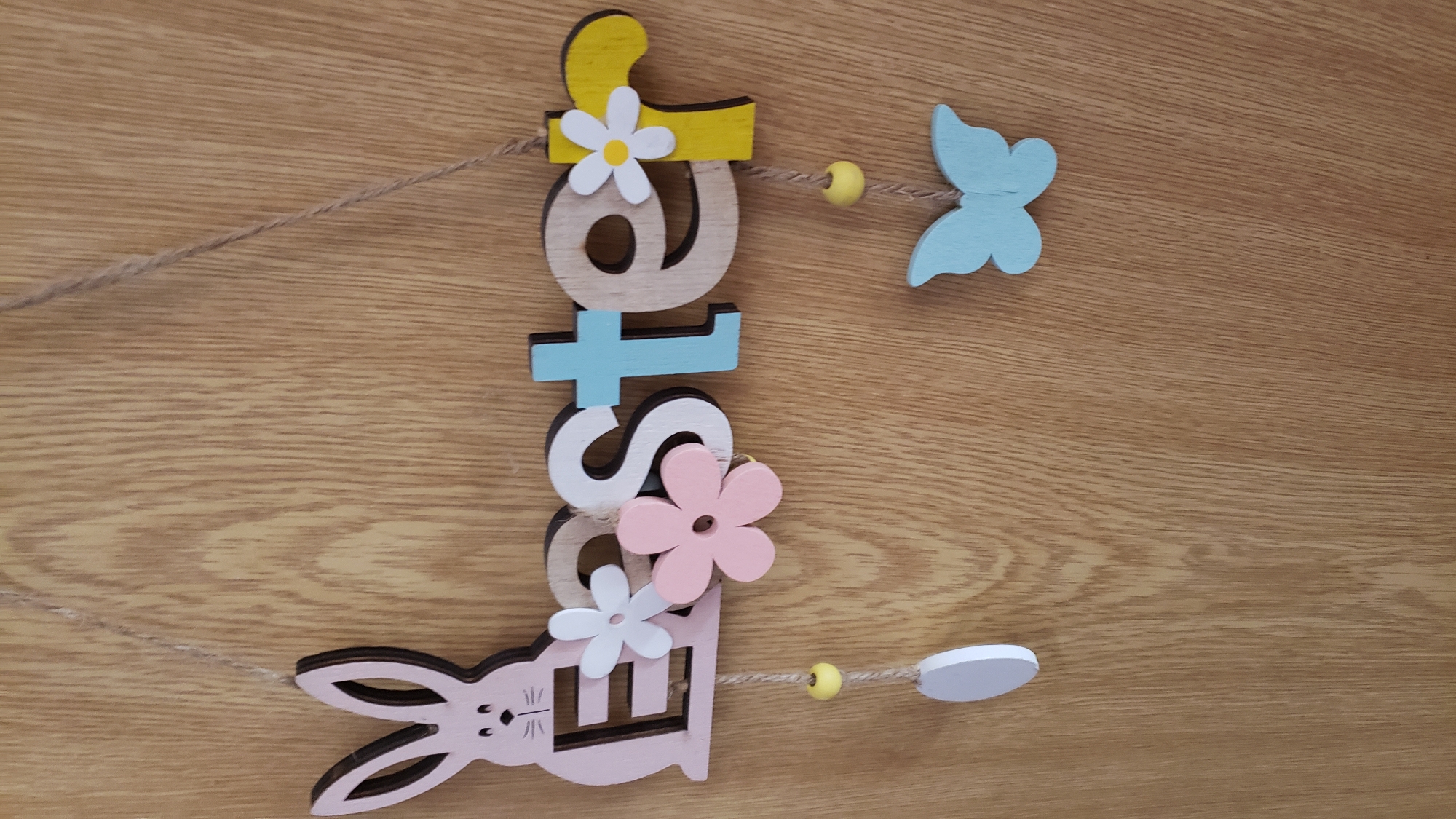 The image shows a wooden Easter decoration hanging on a wooden door or wall. The decoration spells out "Easter" with wooden letters. The "E" is part of a pink Easter bunny silhouette. The letters have various colors and embellishments, including pastel-colored flowers and a light blue butterfly hanging below the word. The wood grain of the door is clearly visible. There are no people or animals present in the image. The setting is indoors, against a light brown wooden background. There is no information available to determine the location within Bratislava, Slovakia.