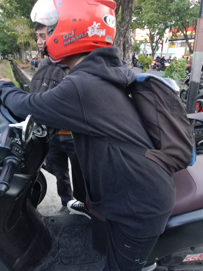The image depicts a man wearing a black hoodie and a red helmet with the words "DJ MARULI" on it, seated on a black scooter. He is wearing black pants and black shoes. The man is holding a large, brown backpack with blue accents. Behind him, another man can be seen wearing a dark jacket and jeans. The image appears to be taken in a city environment, near a road with several other parked motorcycles and scooters. The location of the image, based on the provided coordinates, is Tugu, Indonesia. The background includes green trees and buildings, with a sign in the distance.